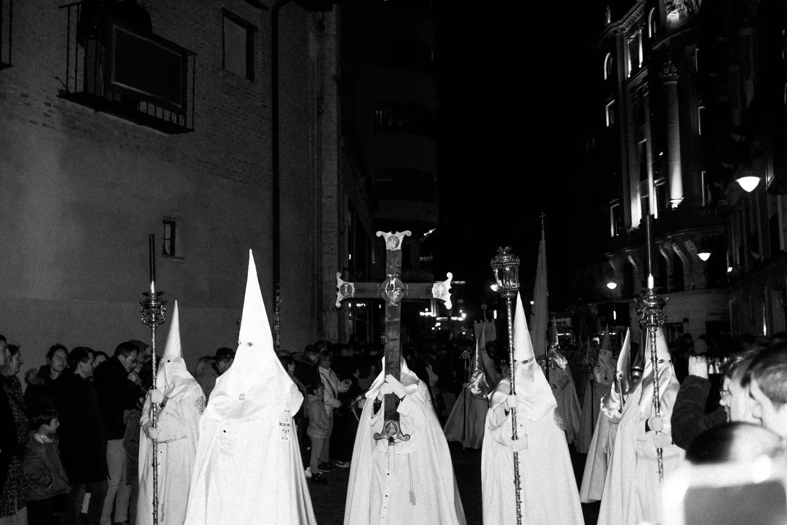 Procesión religiosa en la calle durante la noche, con personas vestidas con túnicas y capuchas blancas, llevando un crucifijo y antorchas, rodeadas de espectadores.