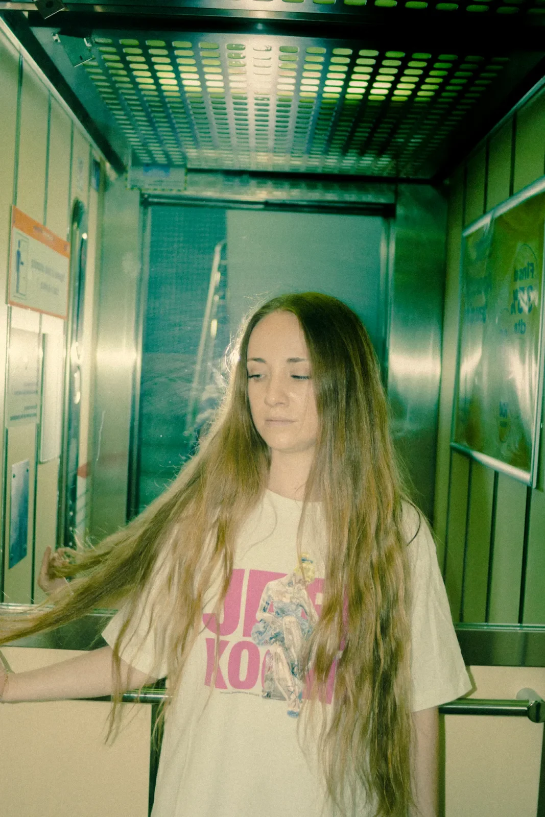 Joven mujer con cabello largo y rubio, usando camiseta beige con ilustración y texto en colores rosa y azul, en un ascensor con paredes metálicas y cartel en la pared a su derecha.