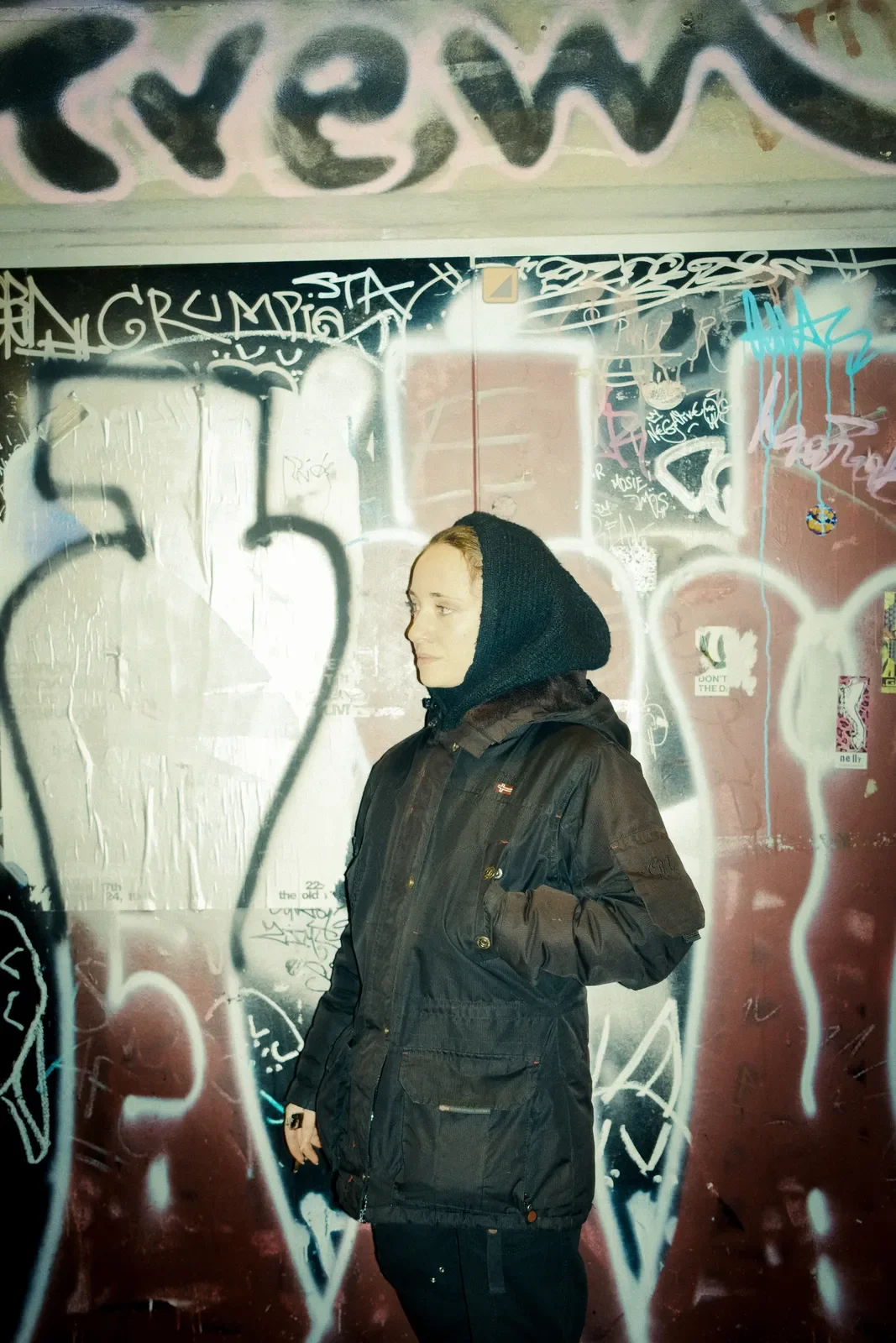 Joven con chaqueta negra y capucha, de perfil, frente a una pared de graffiti en un entorno urbano.