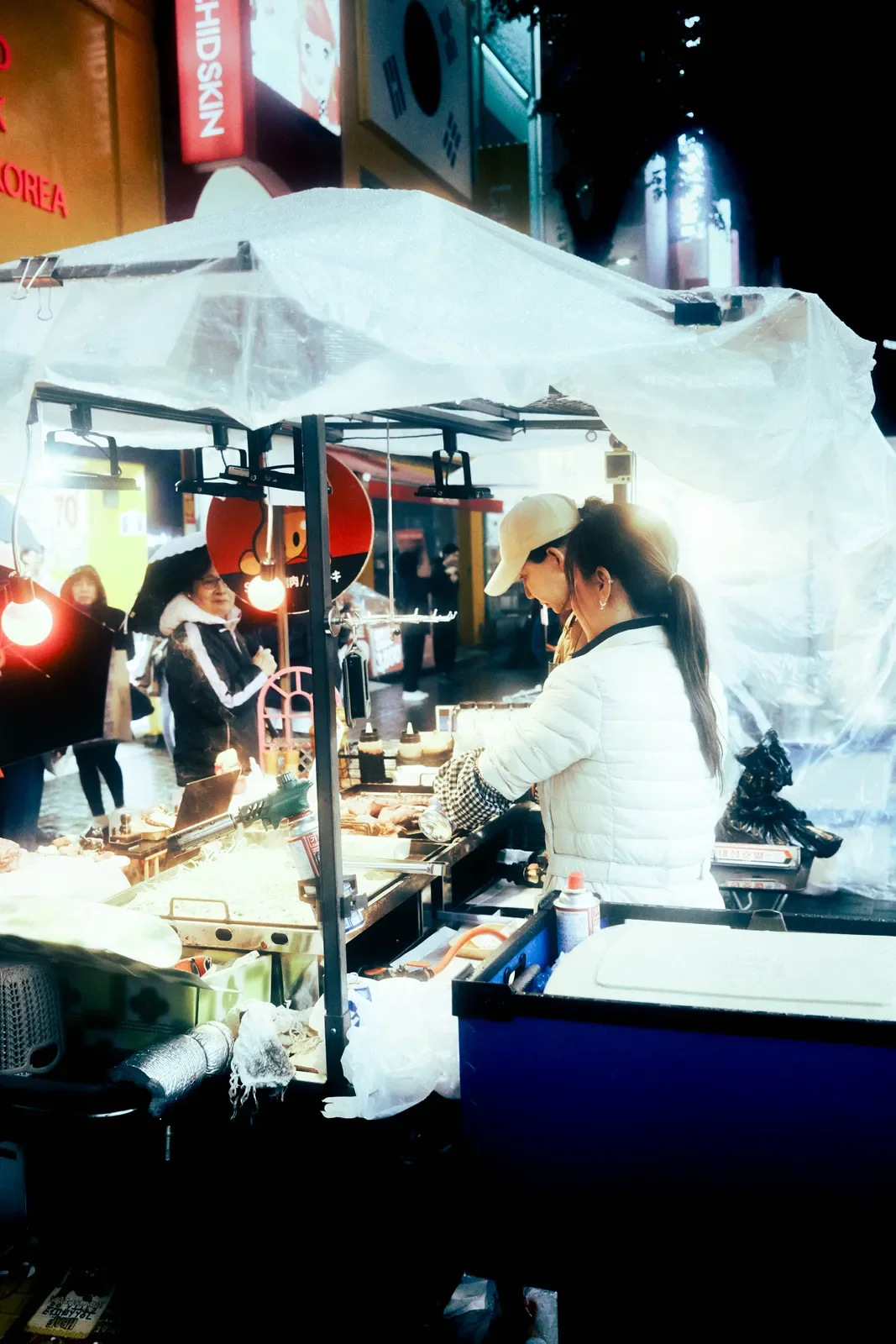 Puestecito callejero con vendedores preparando comida, en noche con iluminación artificial y personas en el fondo.