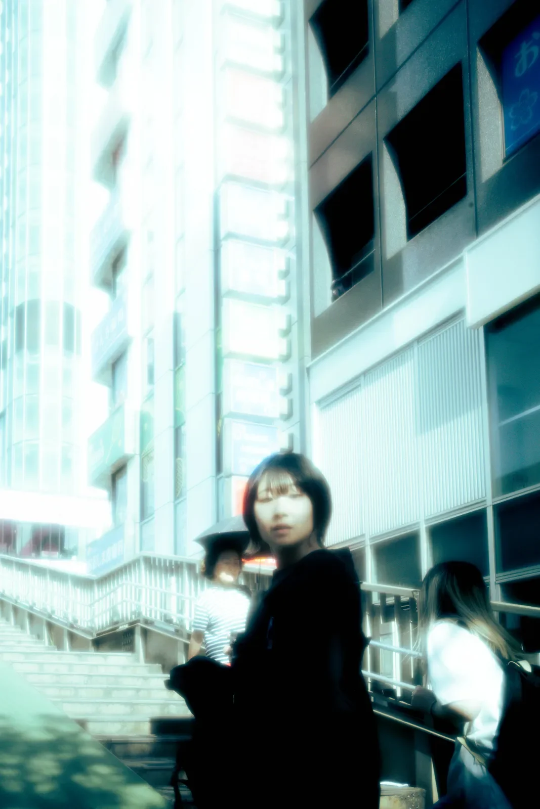 Personas en una ciudad con edificios altos, una de ellas con gafas y cabello corto, mirando hacia la cámara. Algunas personas usan paraguas y están en una escalera.