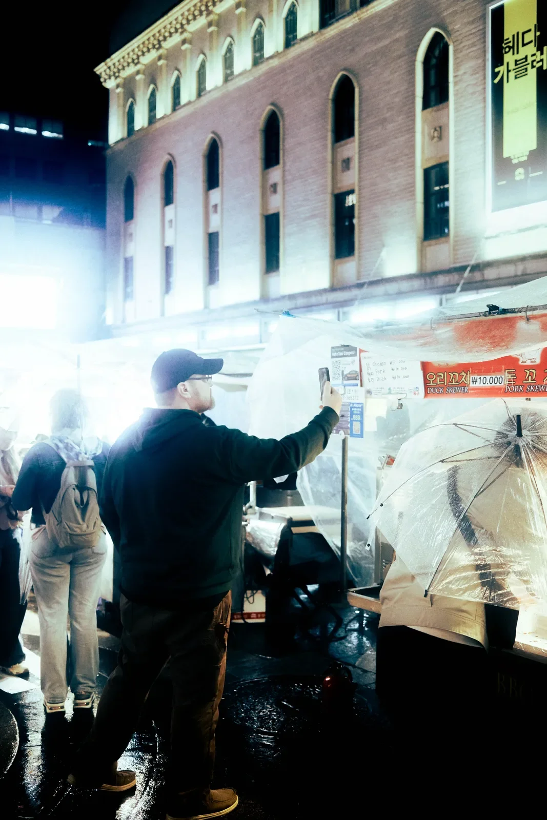 Persona usando un teléfono móvil en un puesto de comida en la calle con paraguas transparente en la noche lluviosa, en una ciudad con edificios antiguos y letreros en diferentes idiomas.