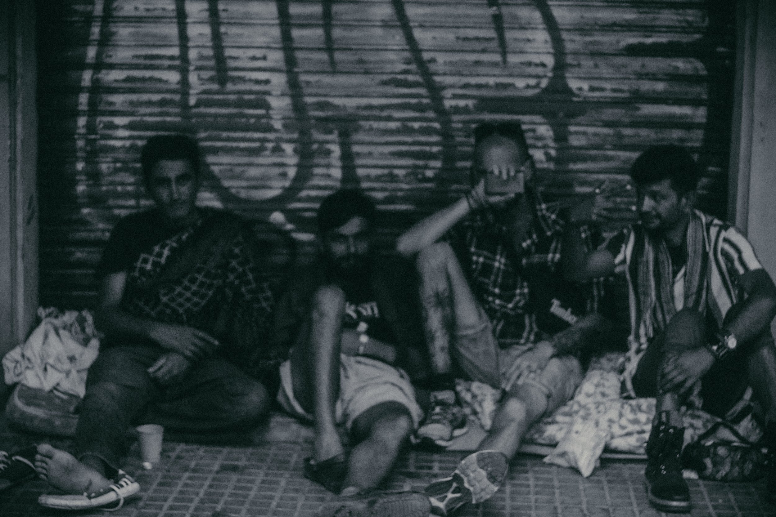 Cuatro personas sentadas en el suelo, con fondo de una persiana de madera, una de ellas se tapa el rostro con la mano. Música y una expresión de tranquilidad en sus rostros.