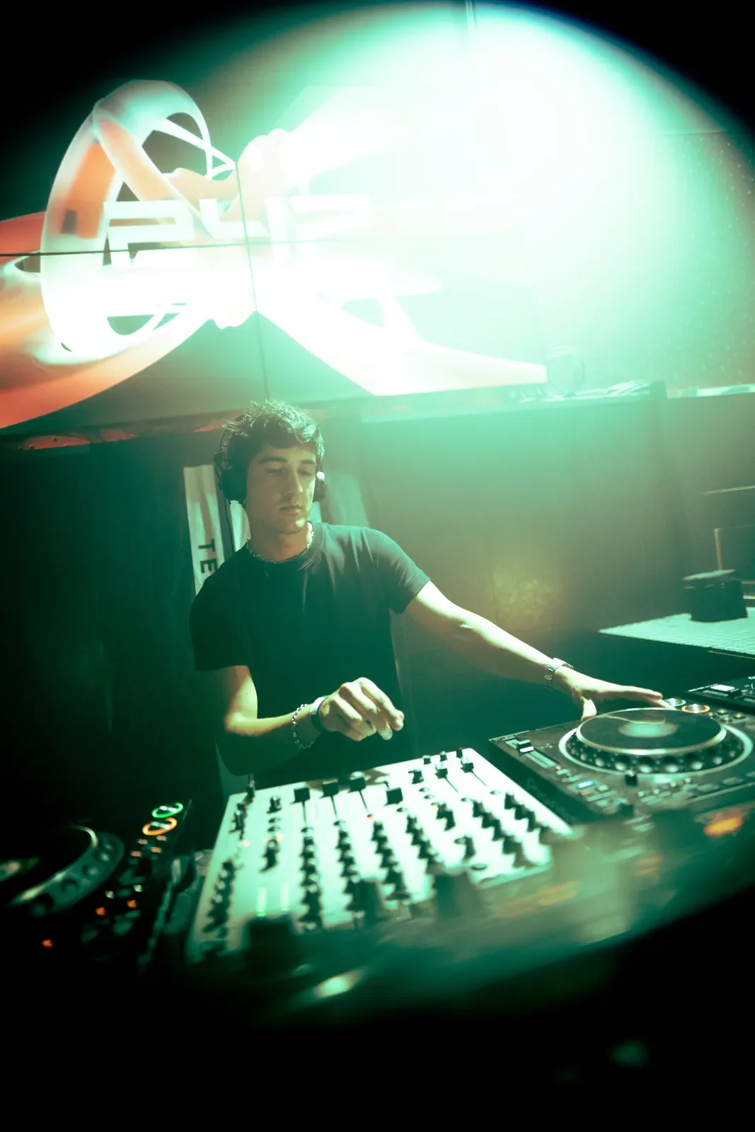 Un DJ tocando en una cabina de discotec con luces verdes y un logo en una pantalla detrás.