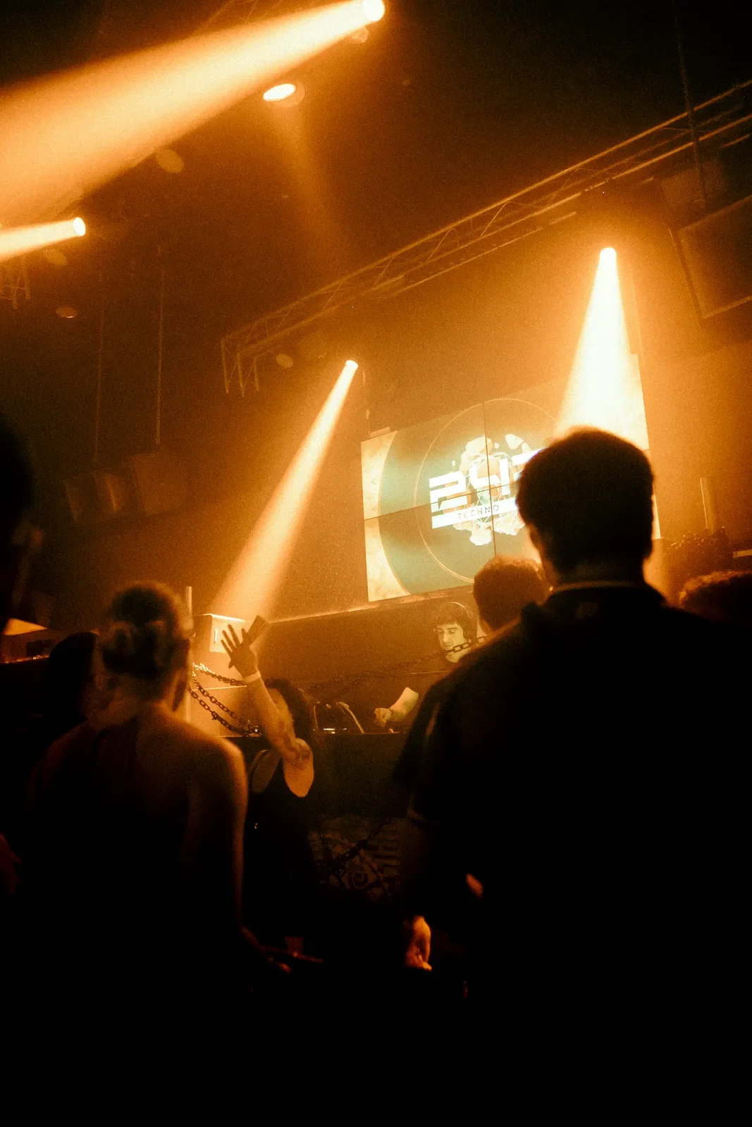 Personas en un club de música electrónica con iluminación tenue y pantalla en el escenario que muestra el logo '2510 Techno'.