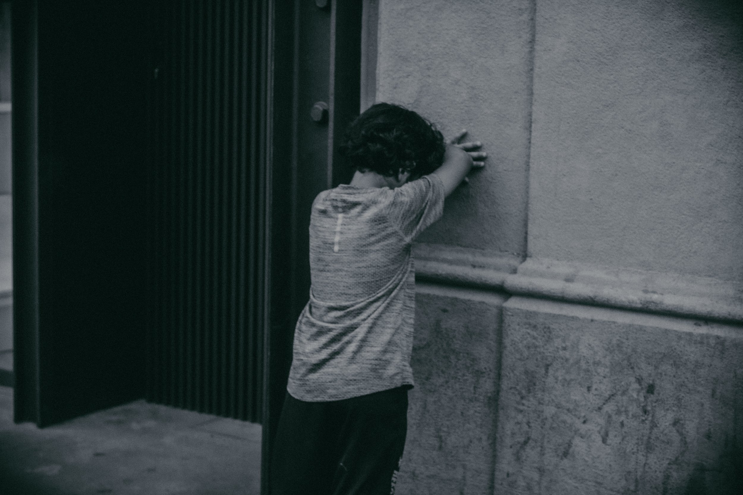Niño con camiseta gris apoya la cabeza y las manos contra una pared de concreto, en un entorno urbano.