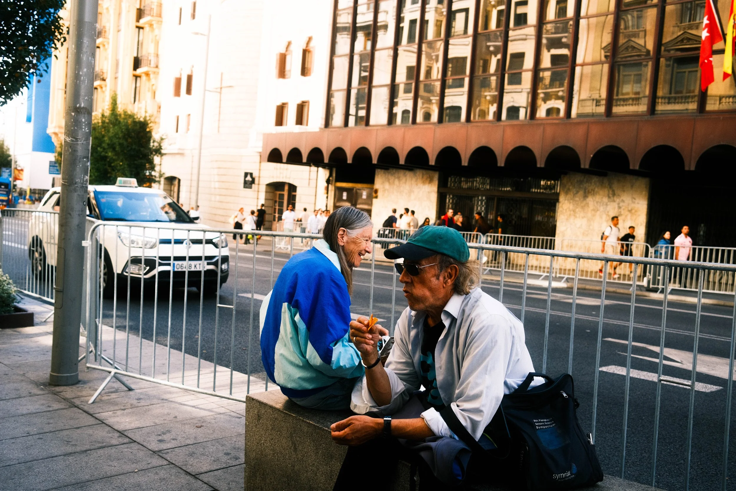 Dos personas sentadas en la acera conversando en la ciudad, con edificios y tráfico en el fondo.