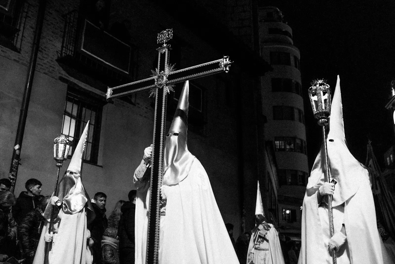 Imagen en blanco y negro de personas realizando una procesión religiosa callejera con personajes con capuchas altos y túnicas, portando un crucifijo y antorchas, en un ambiente nocturno.
