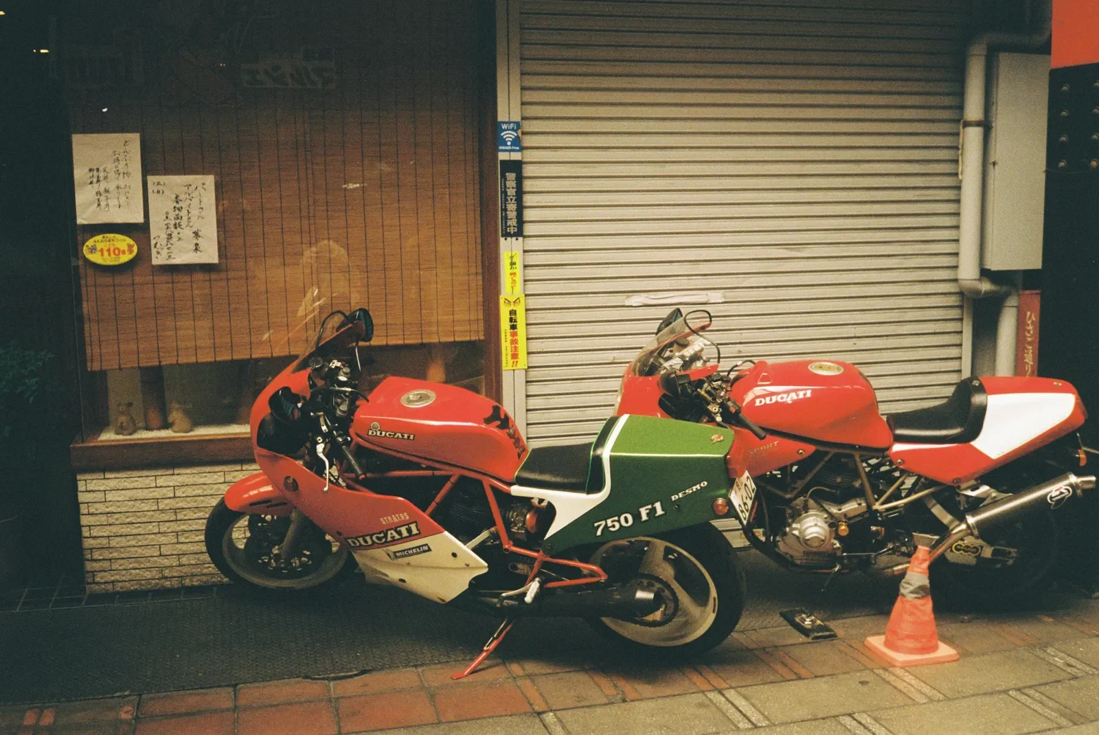 Dos motocicletas Ducati estacionadas en la calle frente a un local cerrado con persiana de metal y ventanas con cortinas. Una de ellas tiene un cono de tráfico junto a la rueda trasera.