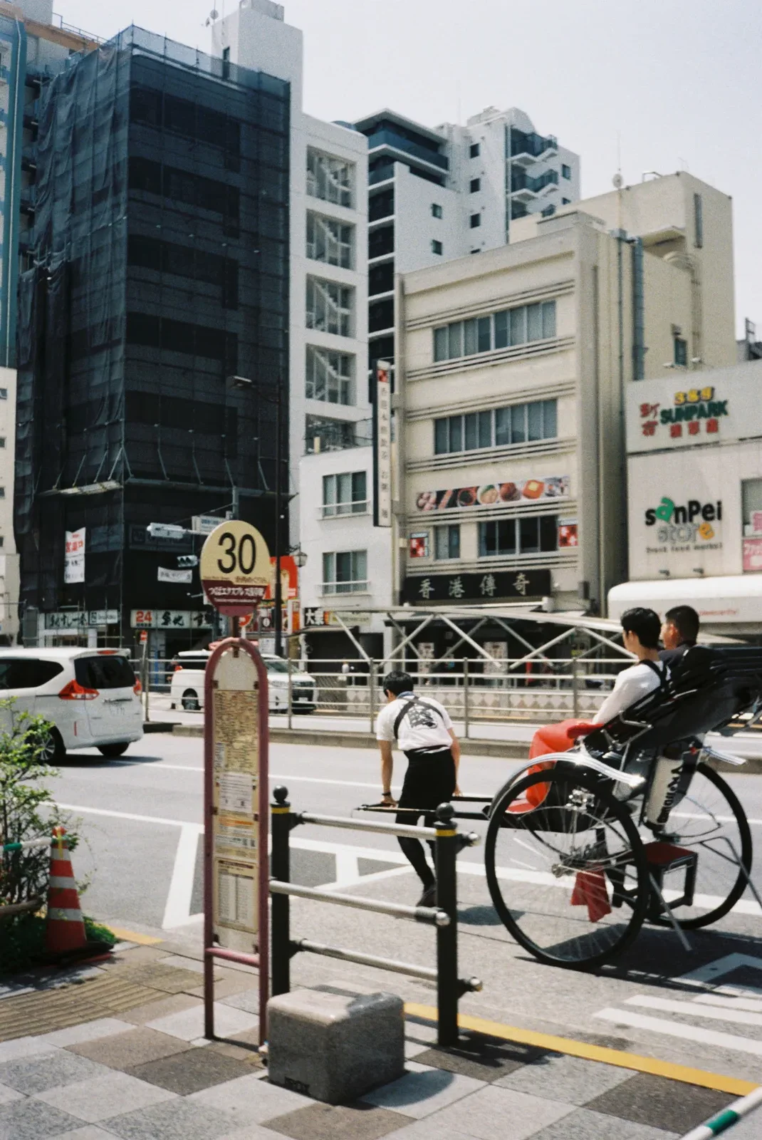 Escena urbana en una ciudad con edificios altos, coches y ciclistas en la calle, señal de velocidad de 30 km/h y señal de tráfico en japonés.