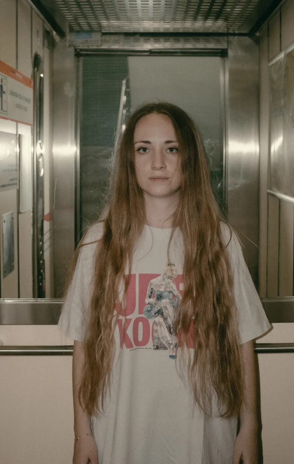Joven con cabello largo y ondulado en un lugar con paredes de metal, usando camiseta con gráfico y letras, de pie y mirando al frente.