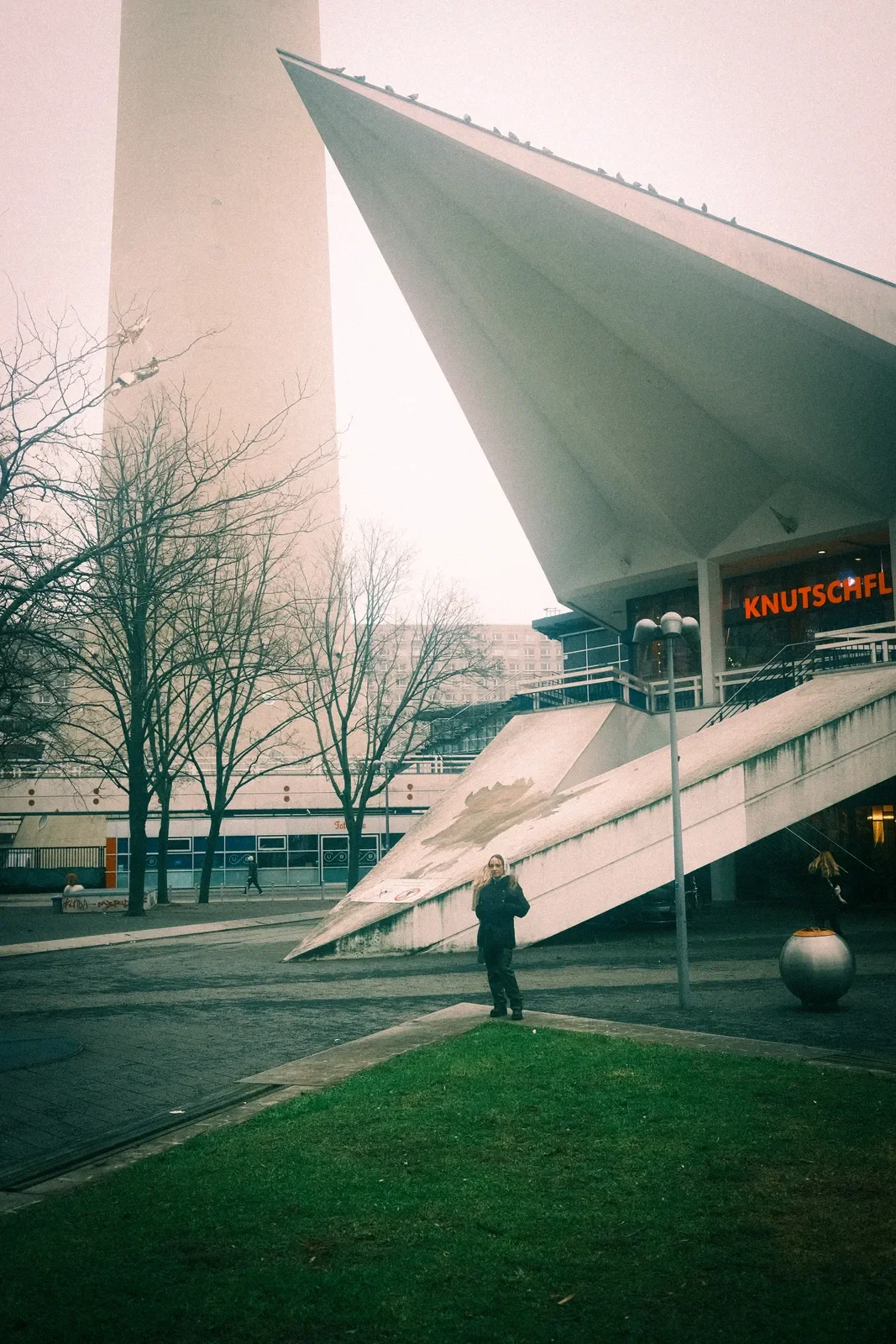 Edificio moderno con estructura angular y letrero de luces rojas que dice 'KNUTSCHFL', árboles sin hojas, persona con abrigo en la acera, área de césped y luz de farola.
