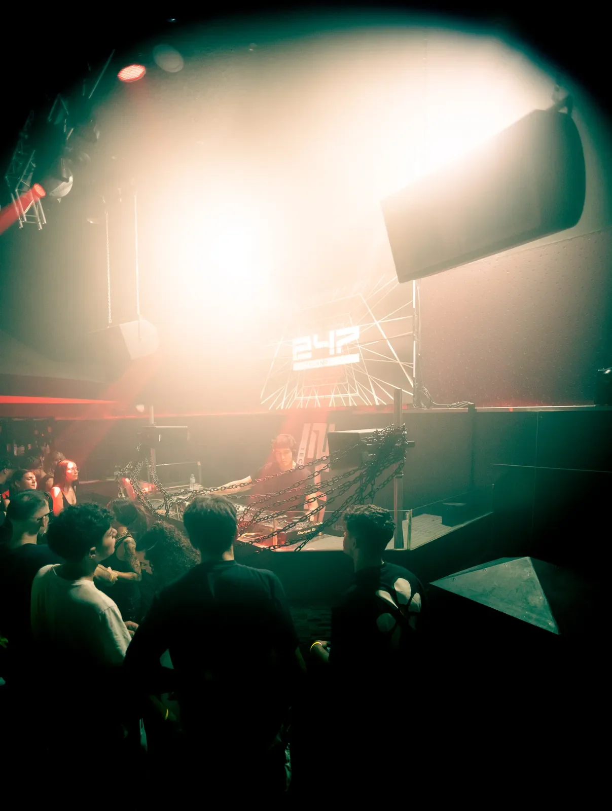 Interior de un club nocturno con un DJ en la cabina, luz brillante y una pantalla que muestra 2:42, con personas bailando y observando.