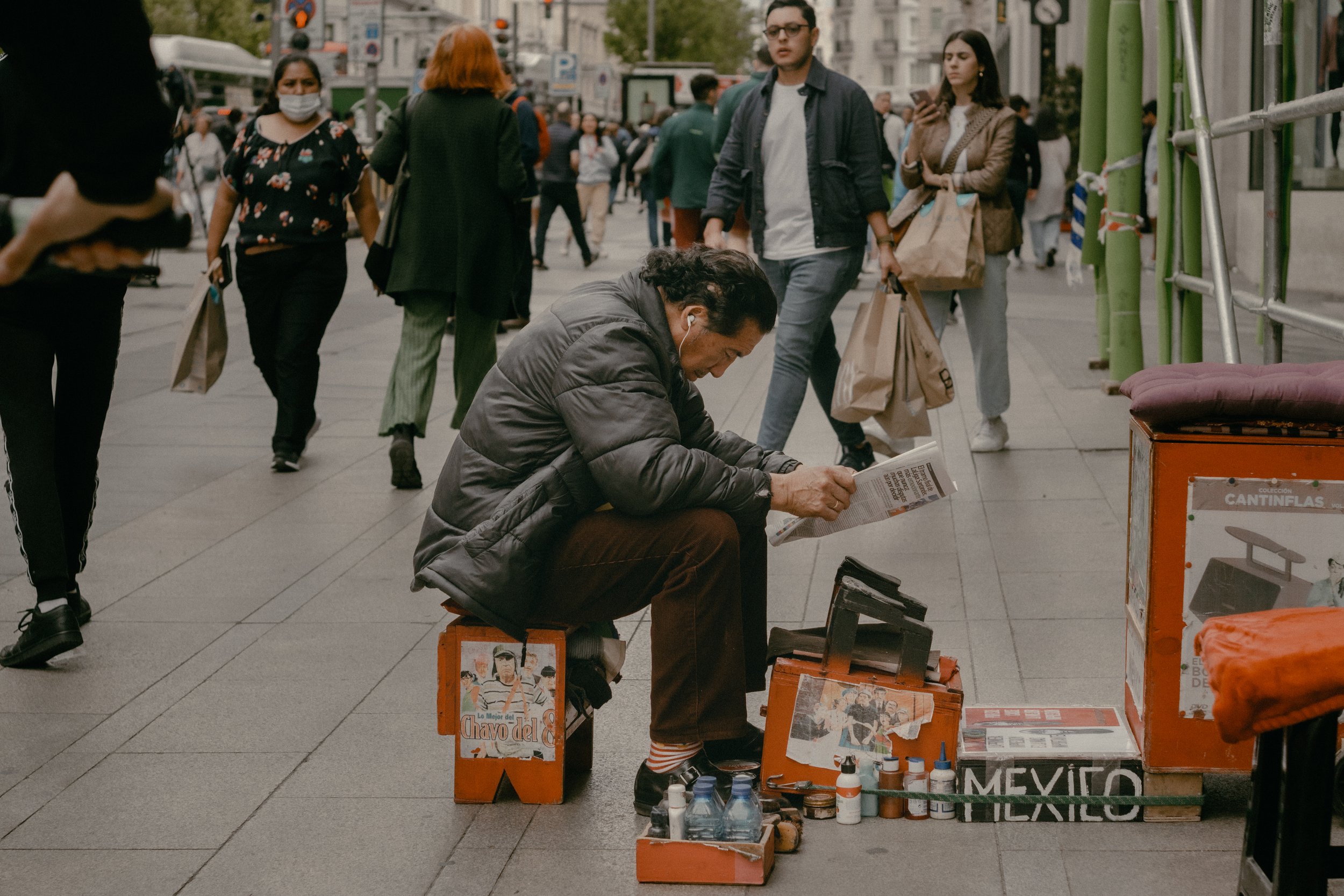 Un hombre sentado en una banca en la acera vendiendo pinturas y objetos. En el fondo, varias personas caminan por la calle, algunas con bolsas en las manos y otras usando mascarillas.