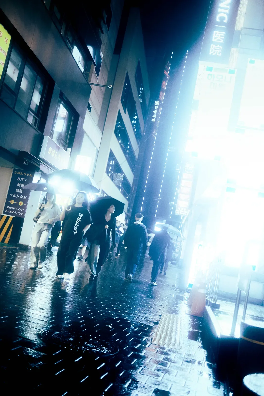 Personas caminando con paraguas en una calle lluviosa en una ciudad moderna durante el día, con edificios altos y anuncios iluminados.