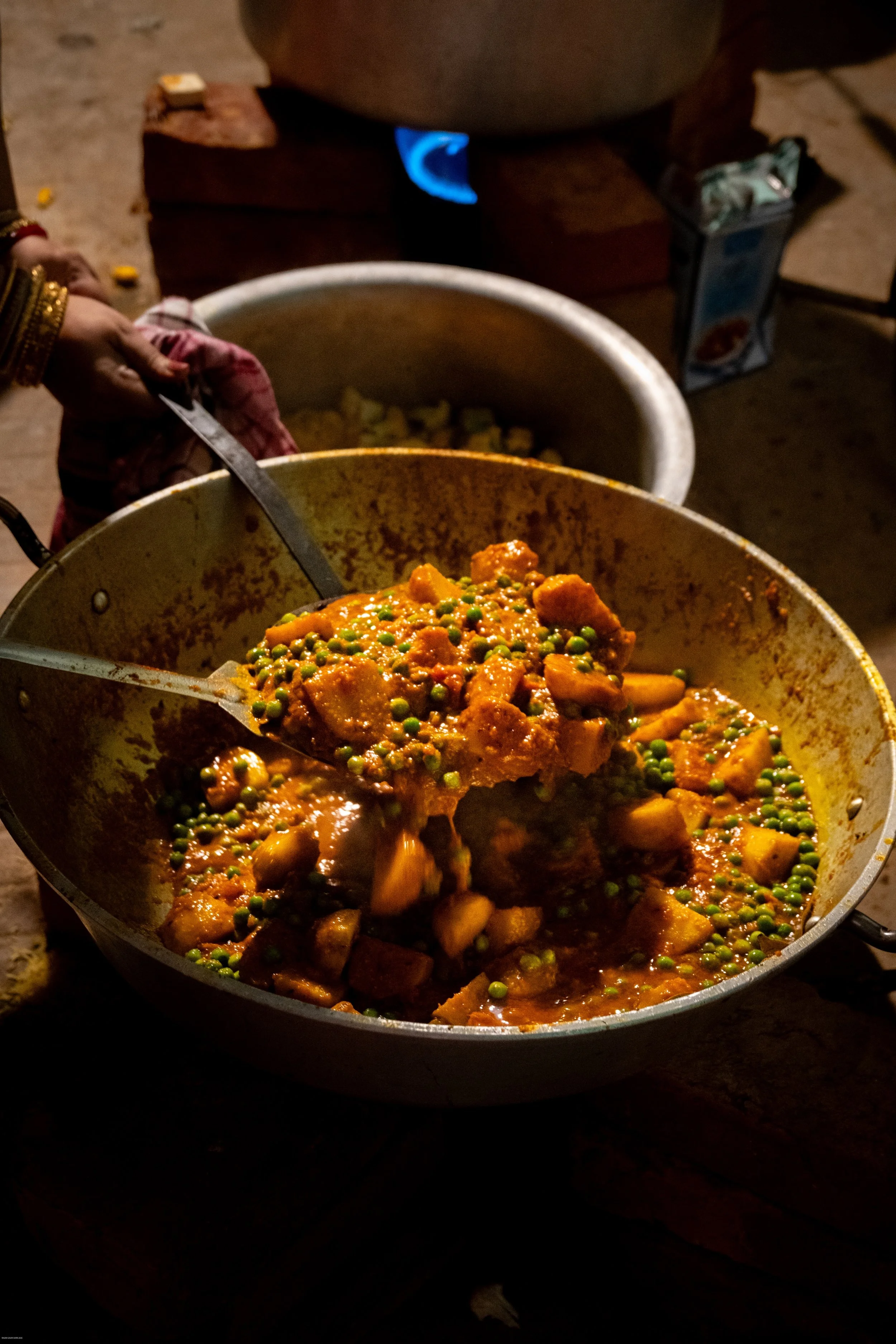 A person serving an Indian vegetable curry with green peas and potatoes from a large cooking pot.