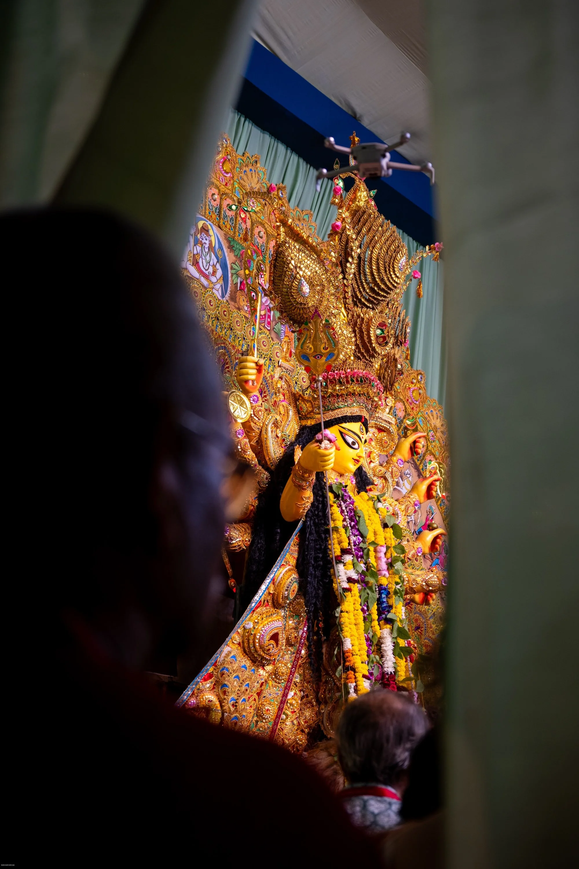 A decorated Hindu deity statue, surrounded by colorful embellishments and flowers, seen through a narrow opening with people in the foreground.