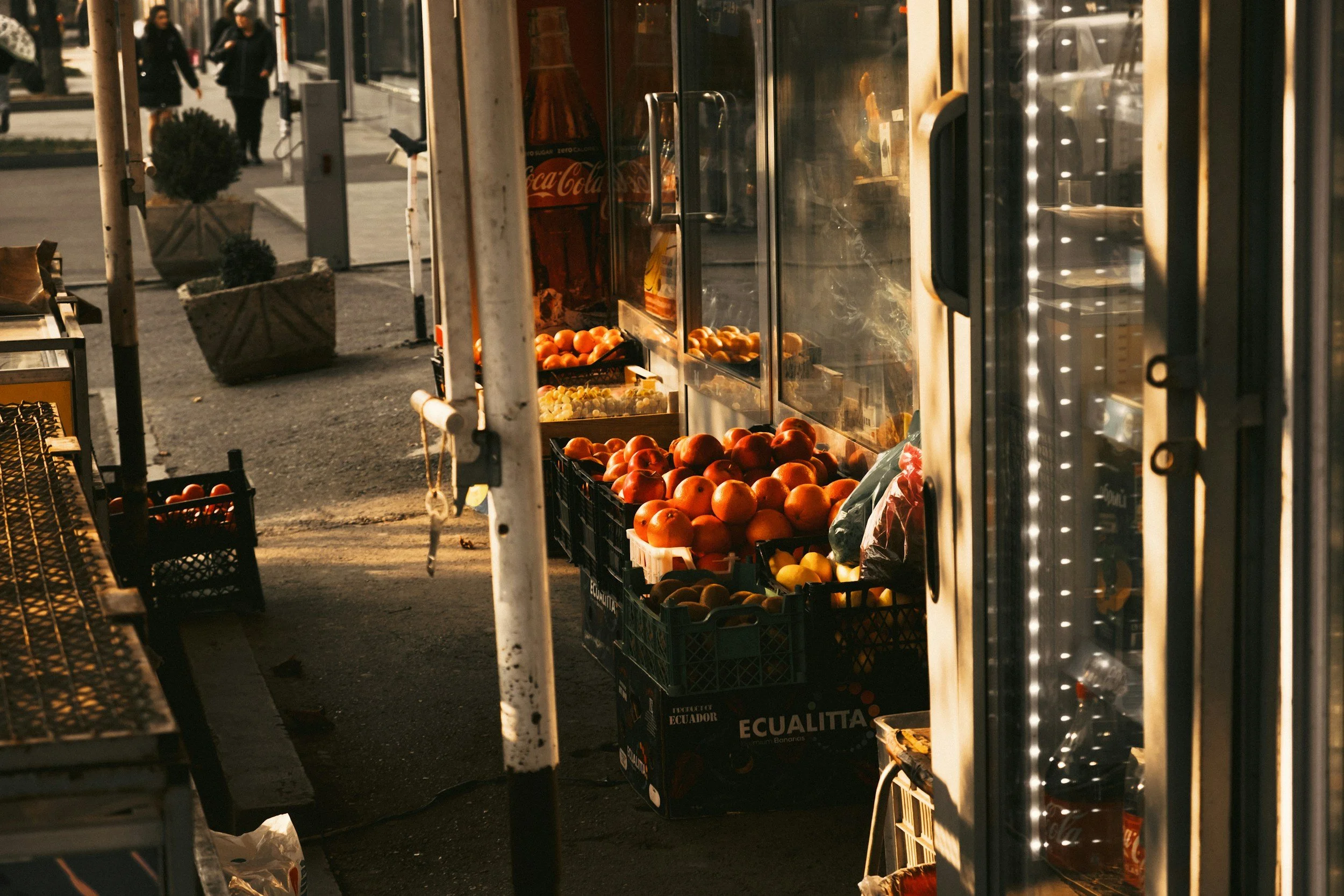 Street vendor stand with fresh fruits, including tomatoes and peaches, illuminated by warm sunlight, in an urban setting.