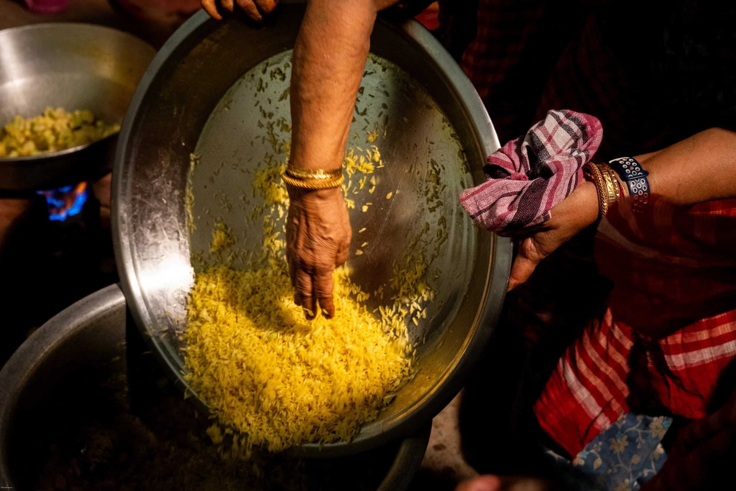 A person with gold bracelets and a pink checkered sleeve is mixing yellow rice in a large stainless steel bowl.