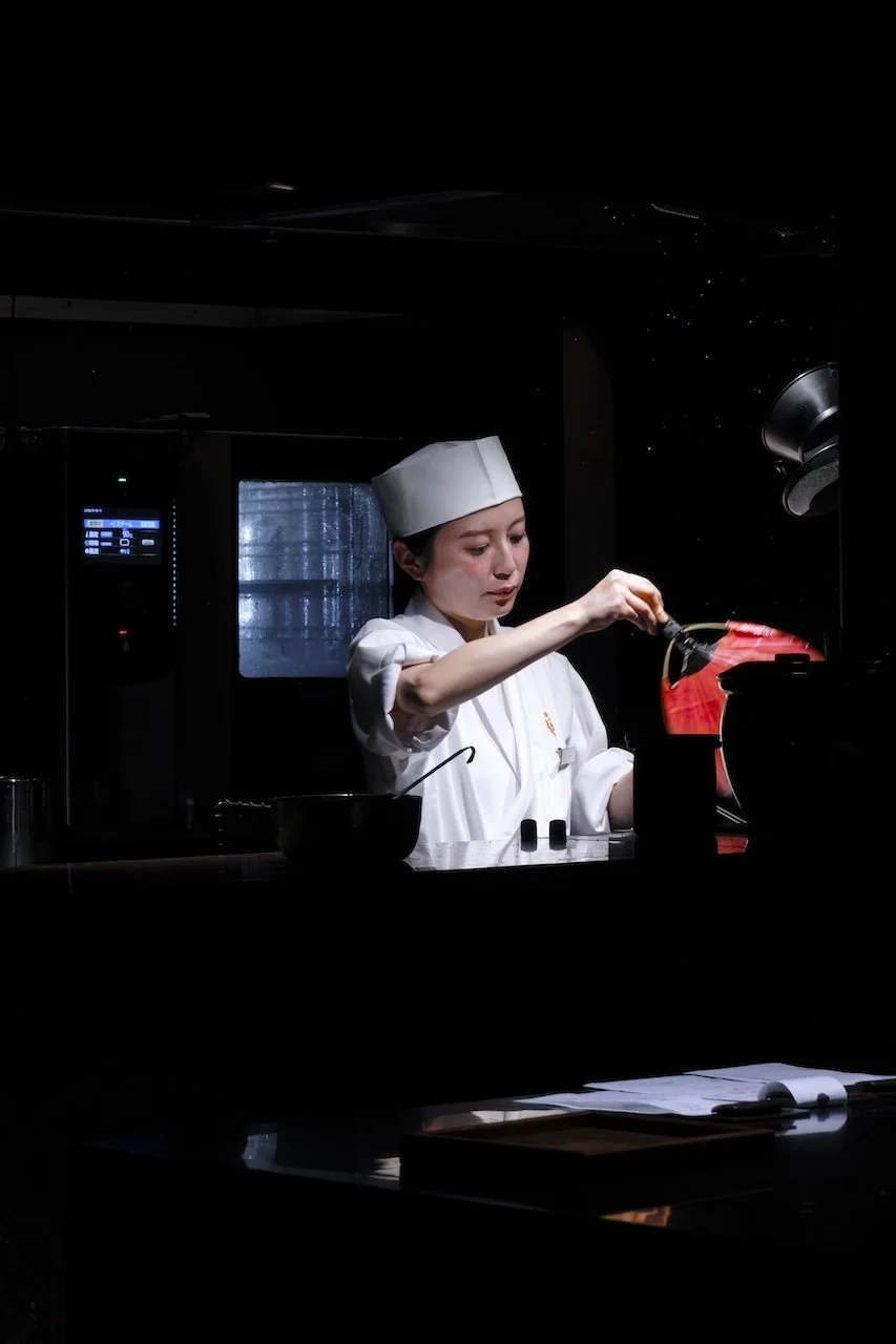 A female chef in a white uniform and hat cooking in a dark kitchen.
