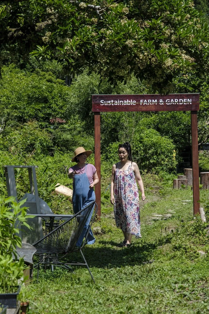 Two women in a green garden near a sign that reads 'Sustainable FARM & GARDEN.' One woman is wearing a sun hat, pink shirt, and overalls, while the other woman is in a floral dress.