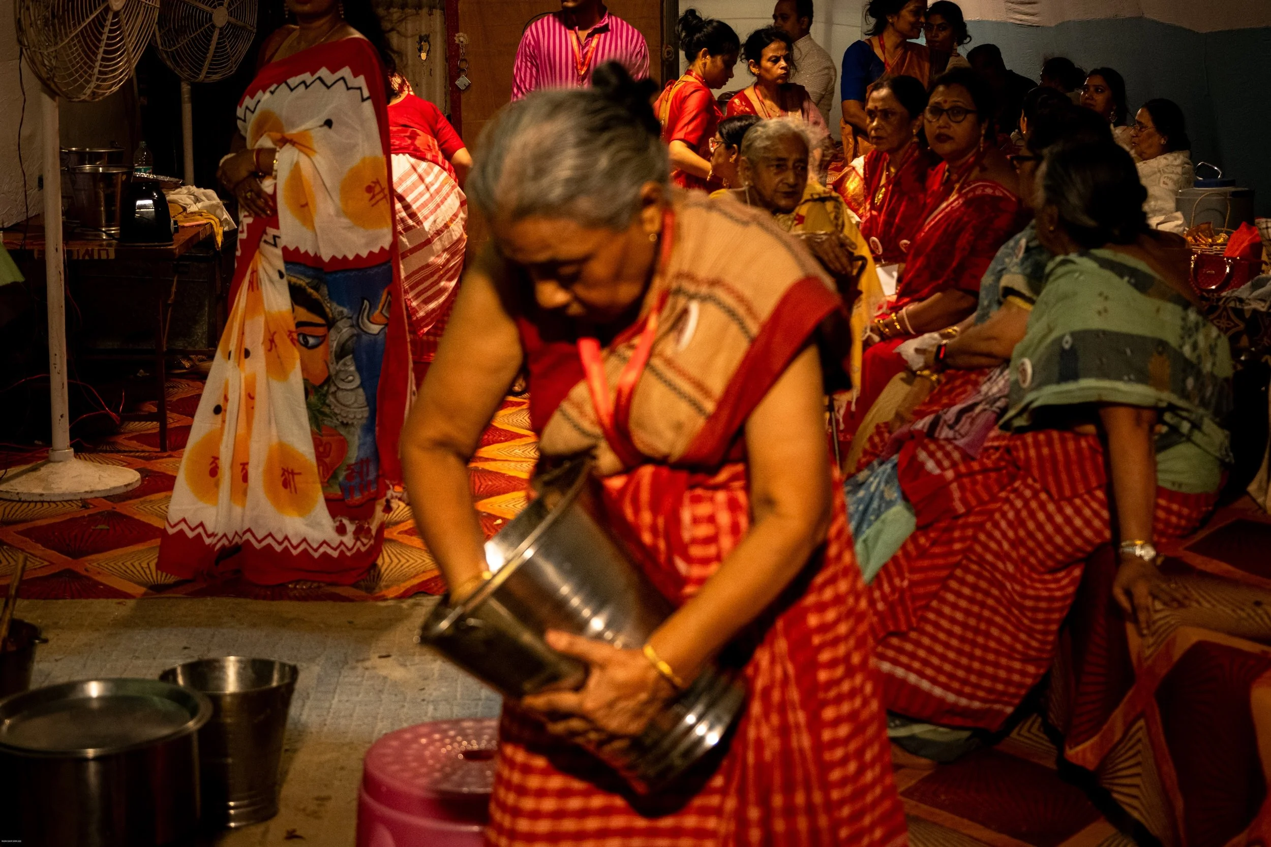 A group of women, mostly elderly, dressed in traditional colorful Indian sarees, gathered in a room. One woman in the foreground is transferring a liquid from a silver container. Others are seated or standing in the background, engaged in conversatio