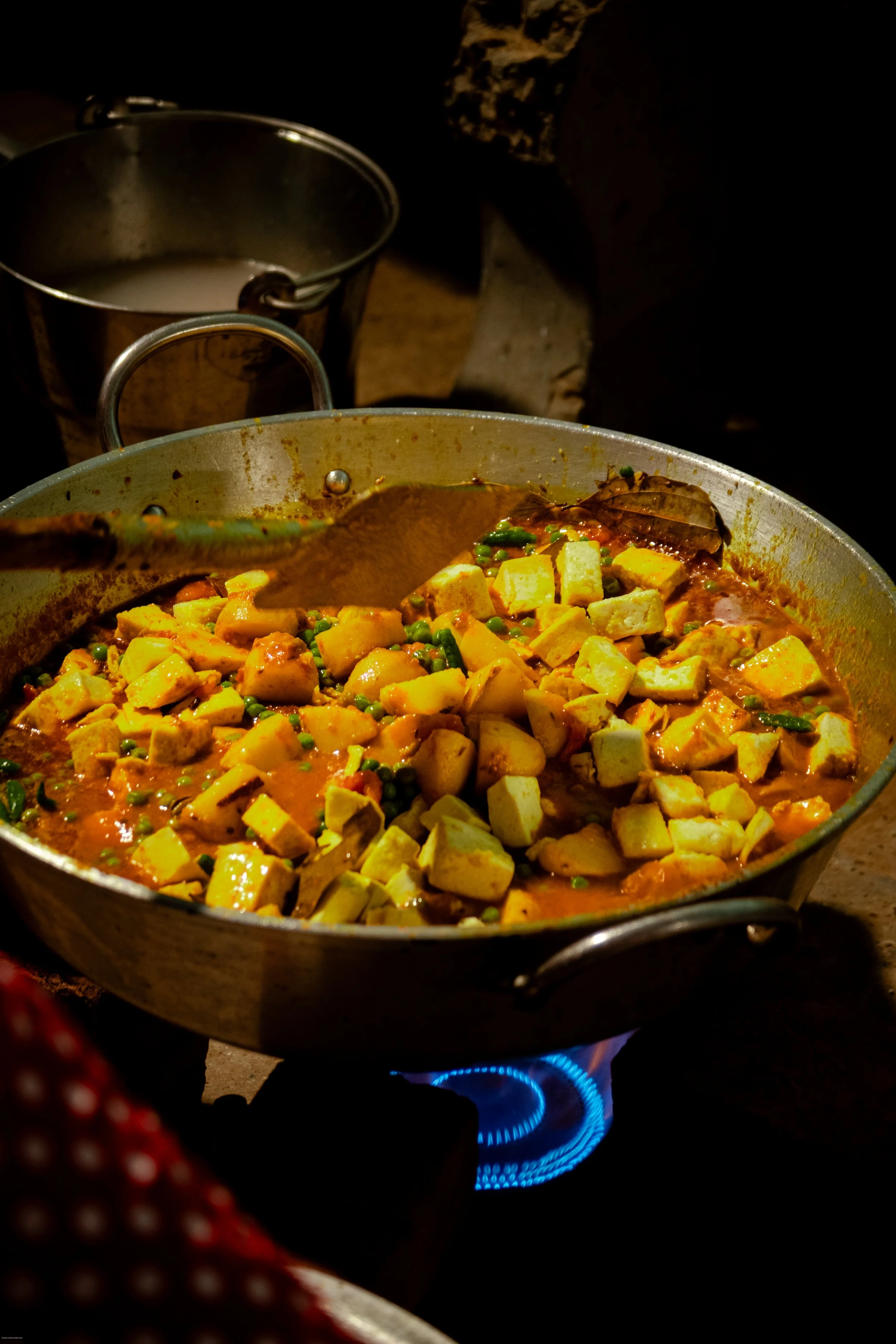 A large pan of tofu and vegetable curry cooking on a stove with a visible blue flame, with a metal spoon inside and a glass of water or milk in the background.