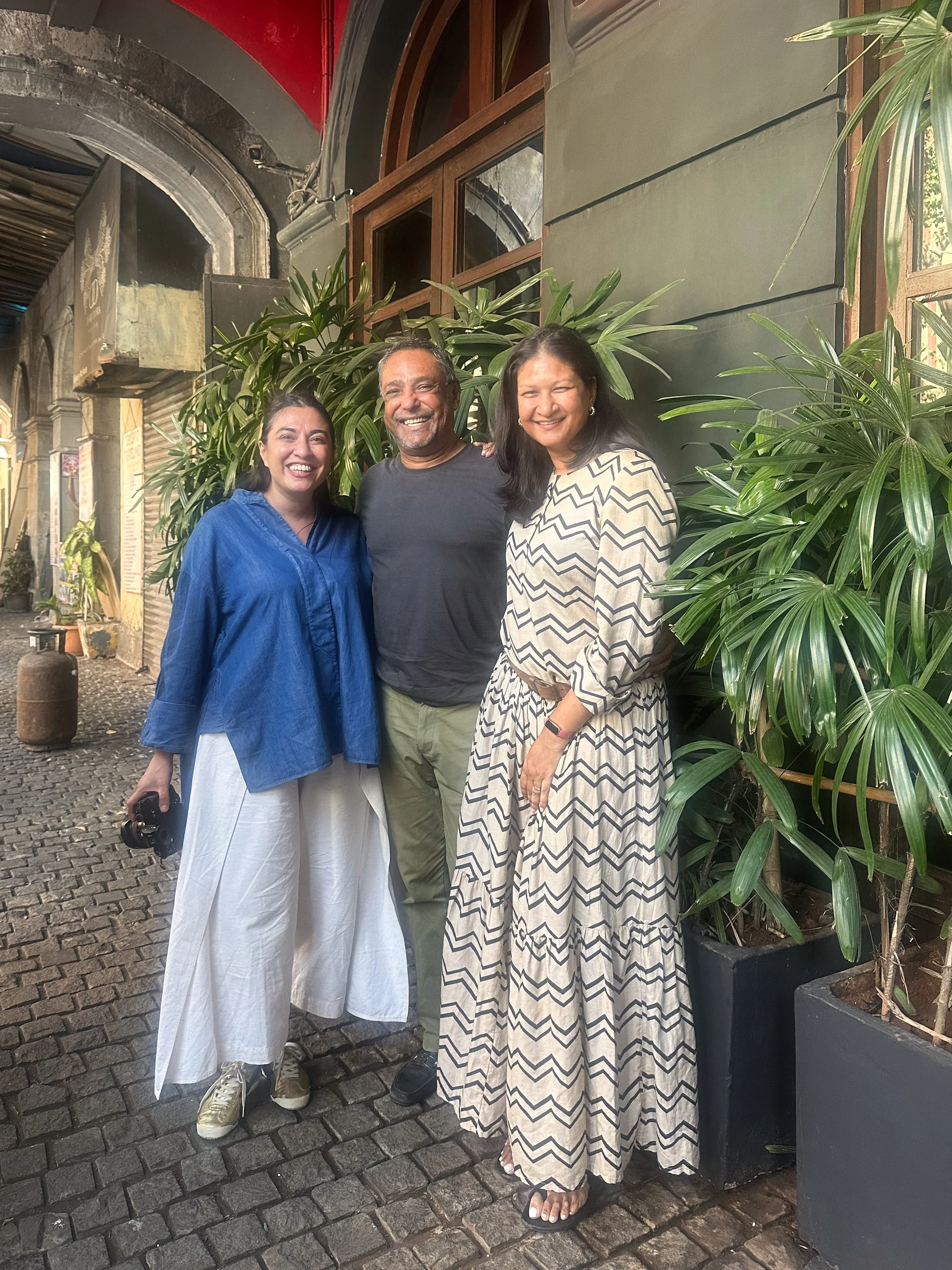 Three people standing close together and smiling outside next to green plants on a cobblestone path.