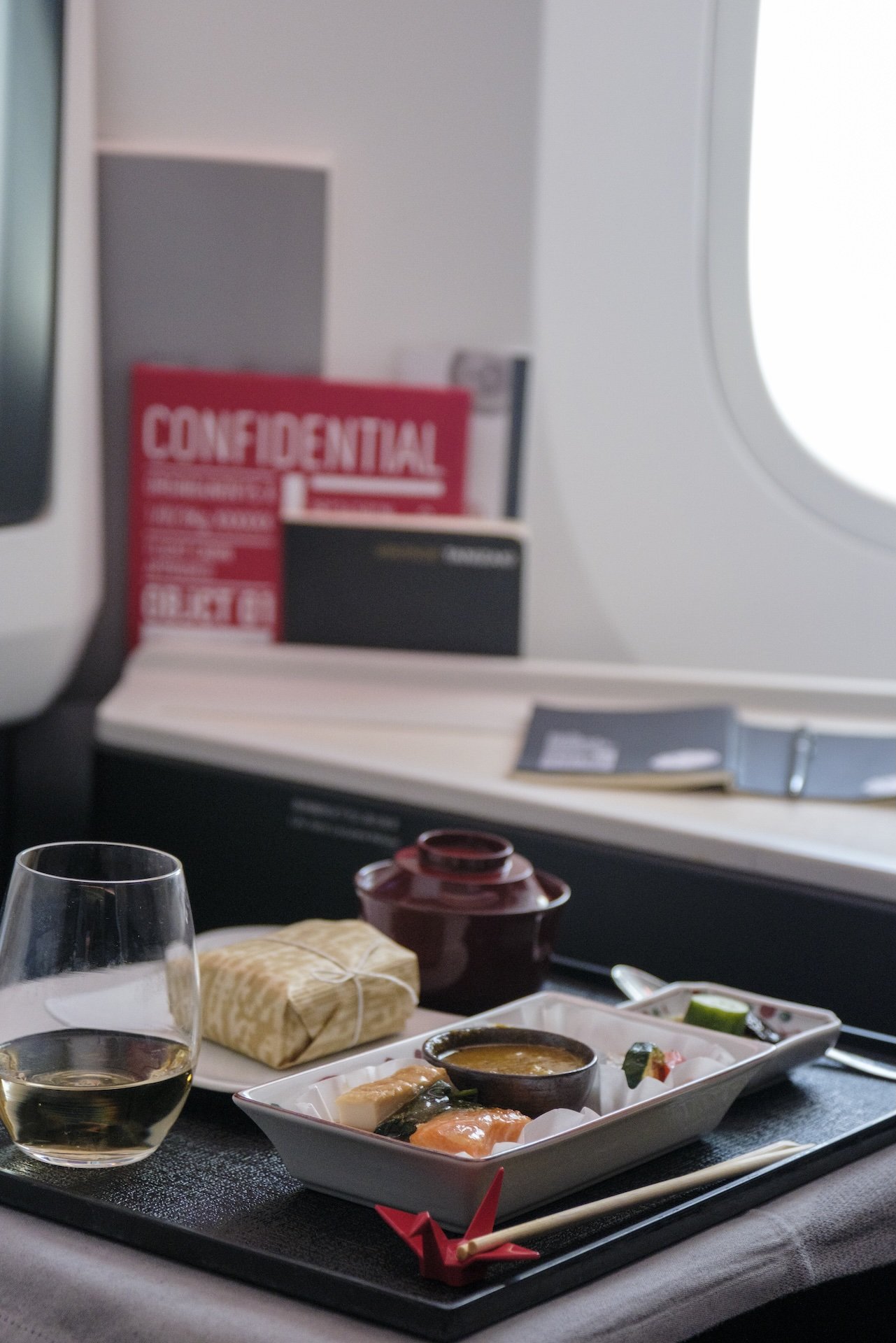 A meal on a tray with sashimi, a small bowl, and a drink, inside an airplane cabin with a window and a safety information card in the background.