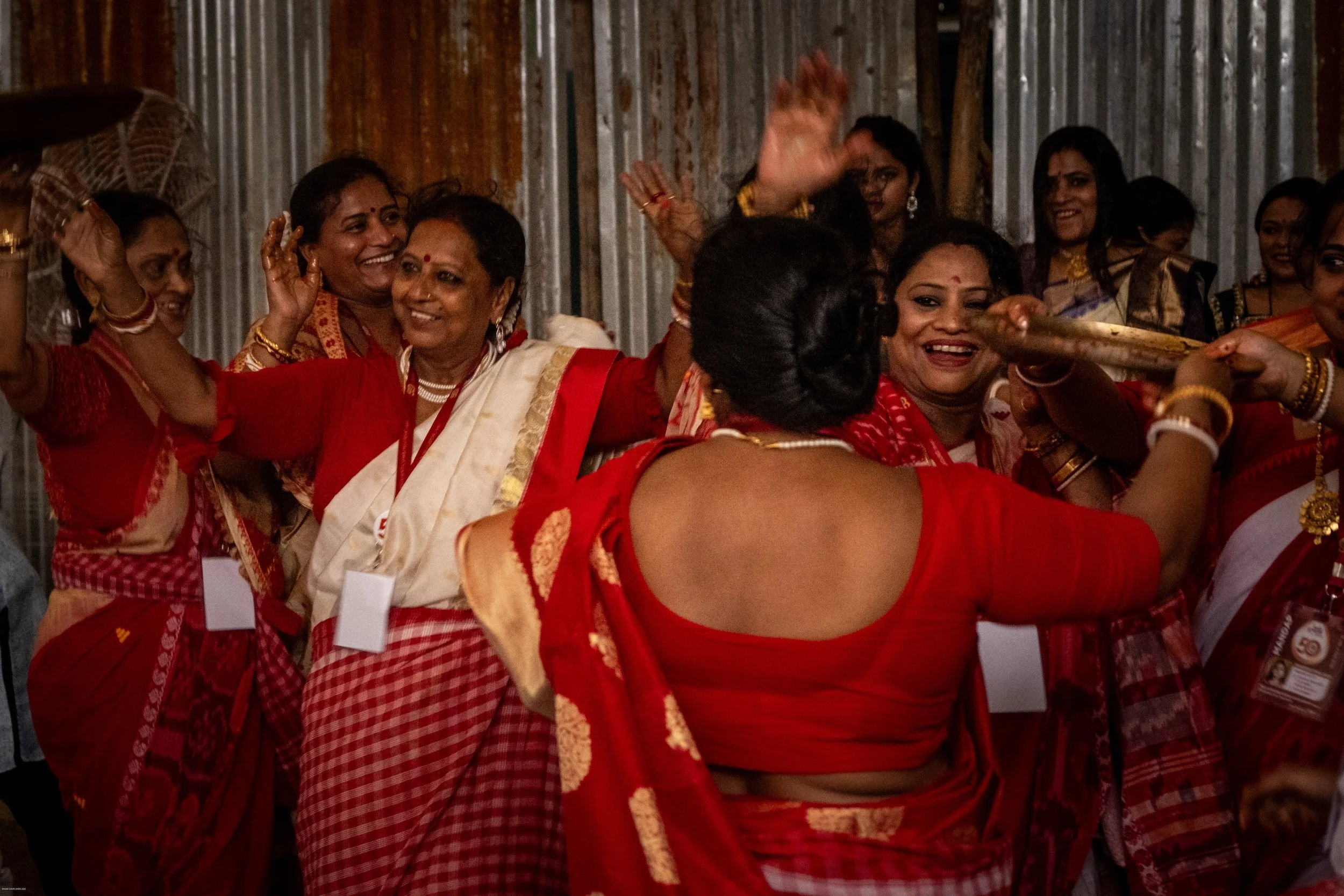 Group of women dressed in traditional red and gold attire celebrating together, some dancing and smiling, in a festive setting.