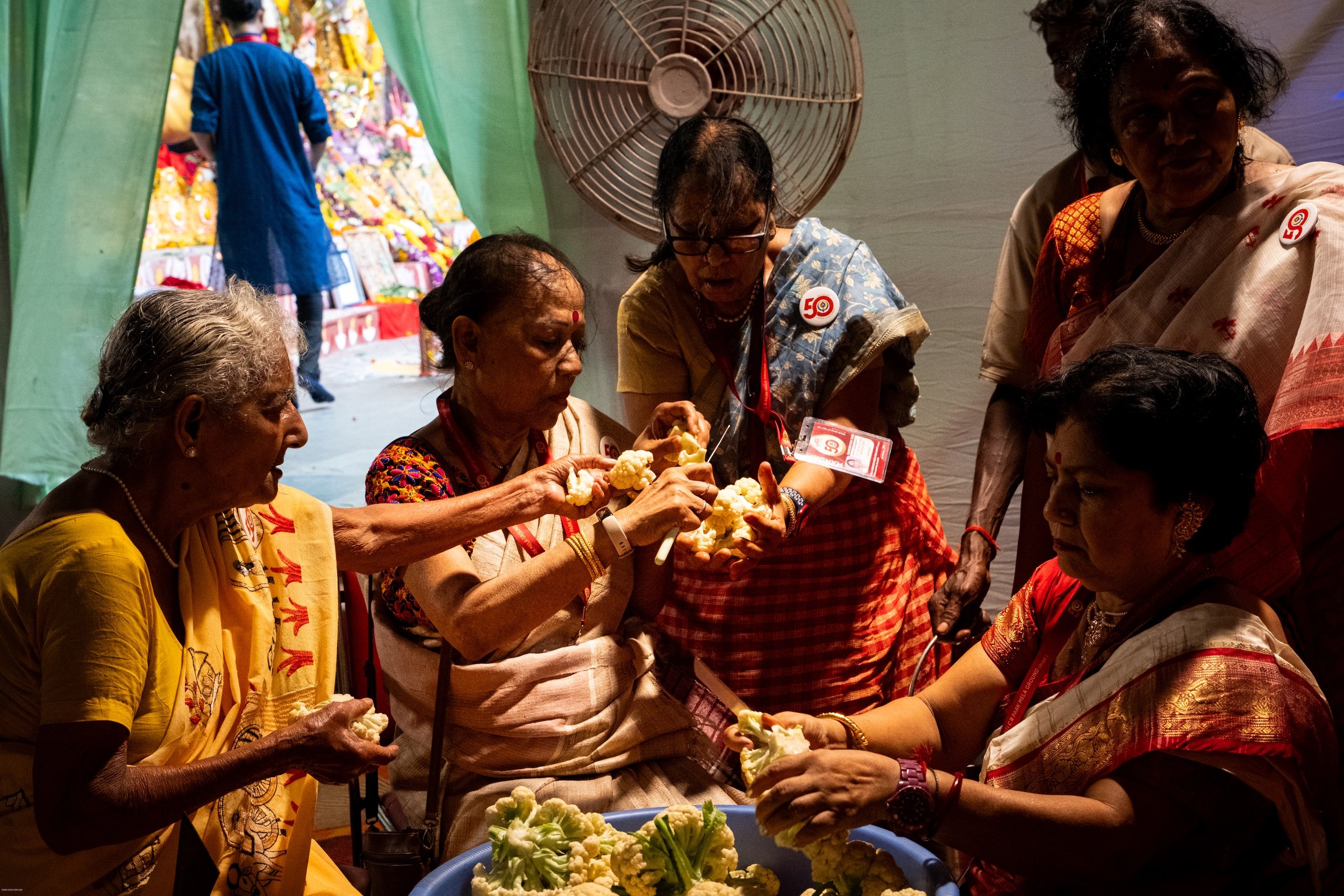 Group of Indian women in traditional attire inspecting cauliflower at a cultural event, with a man in a blue kurta in the background.