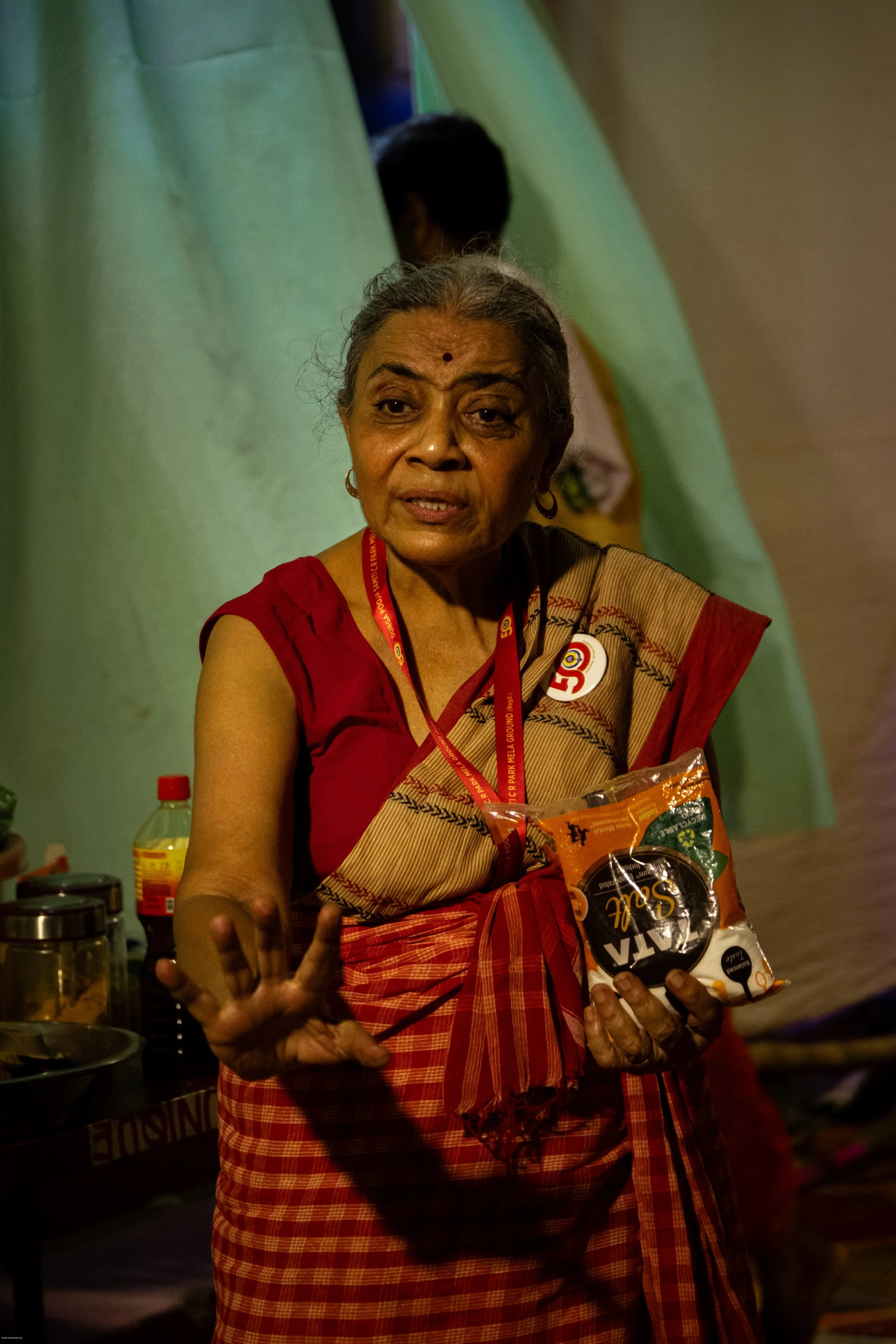 An elderly woman wearing a red saree and a badge with the letter 'G' is holding a packet of snacks and gesturing with her right hand. She appears to be speaking or explaining something at an indoor event.