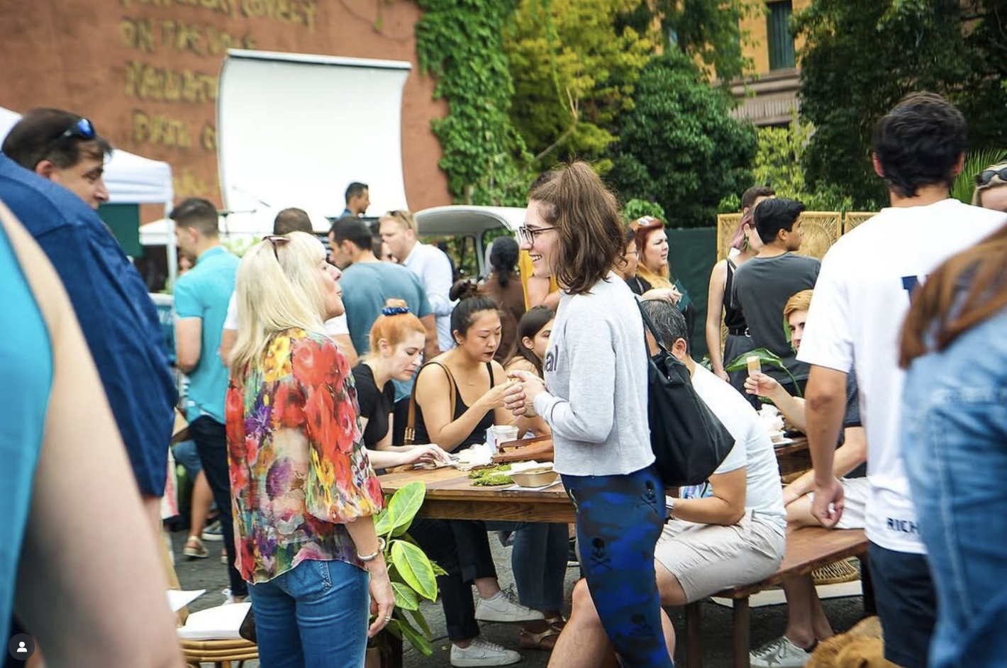 A group of people socializing outdoors at a daytime event, with some sitting at a long wooden table and others standing around, under trees and at a venue with a brick wall in the background.