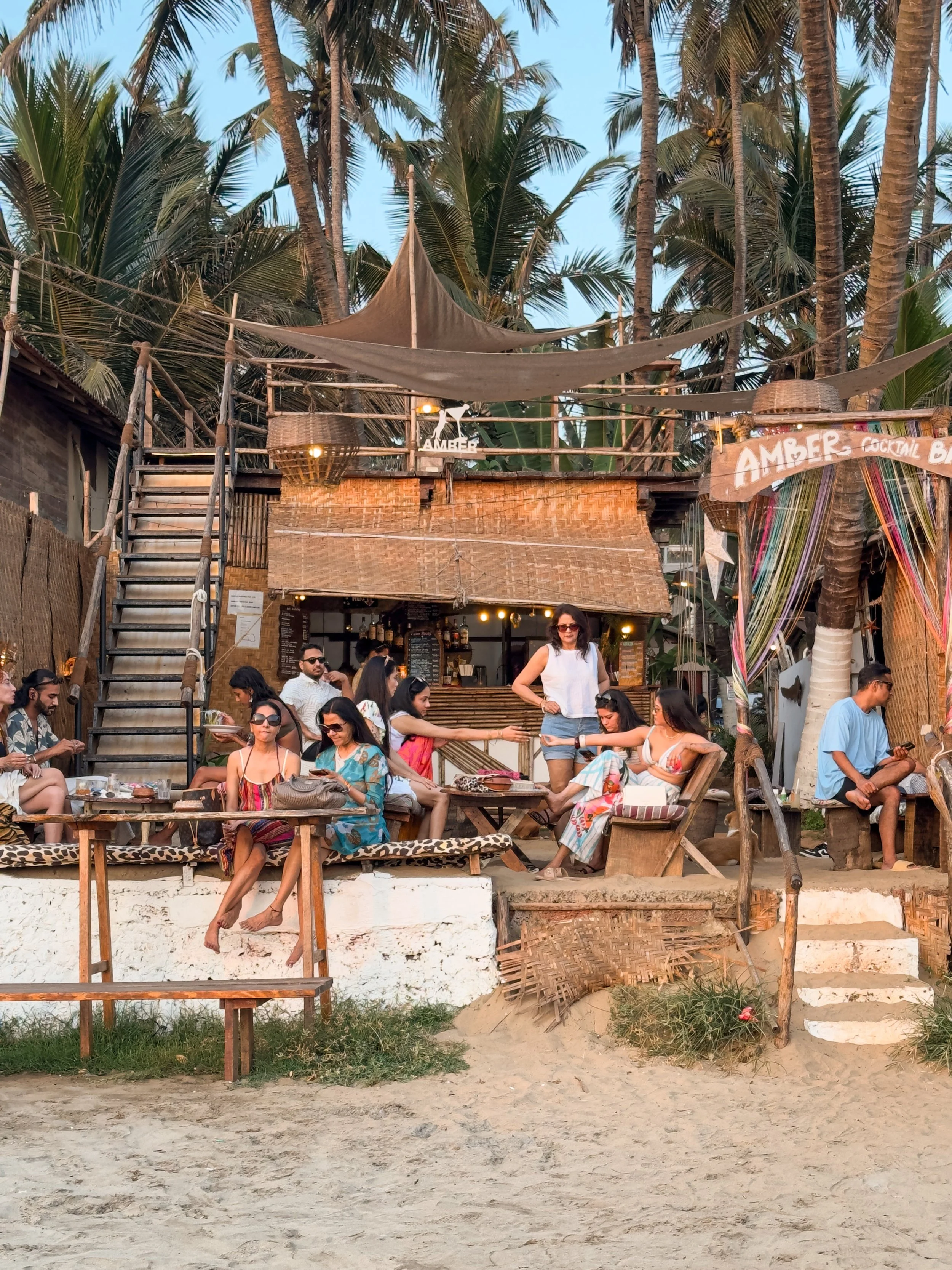 People sitting and socializing outside a beachside bar named Amber, with palm trees, sandy ground, and wooden decor, during sunset.