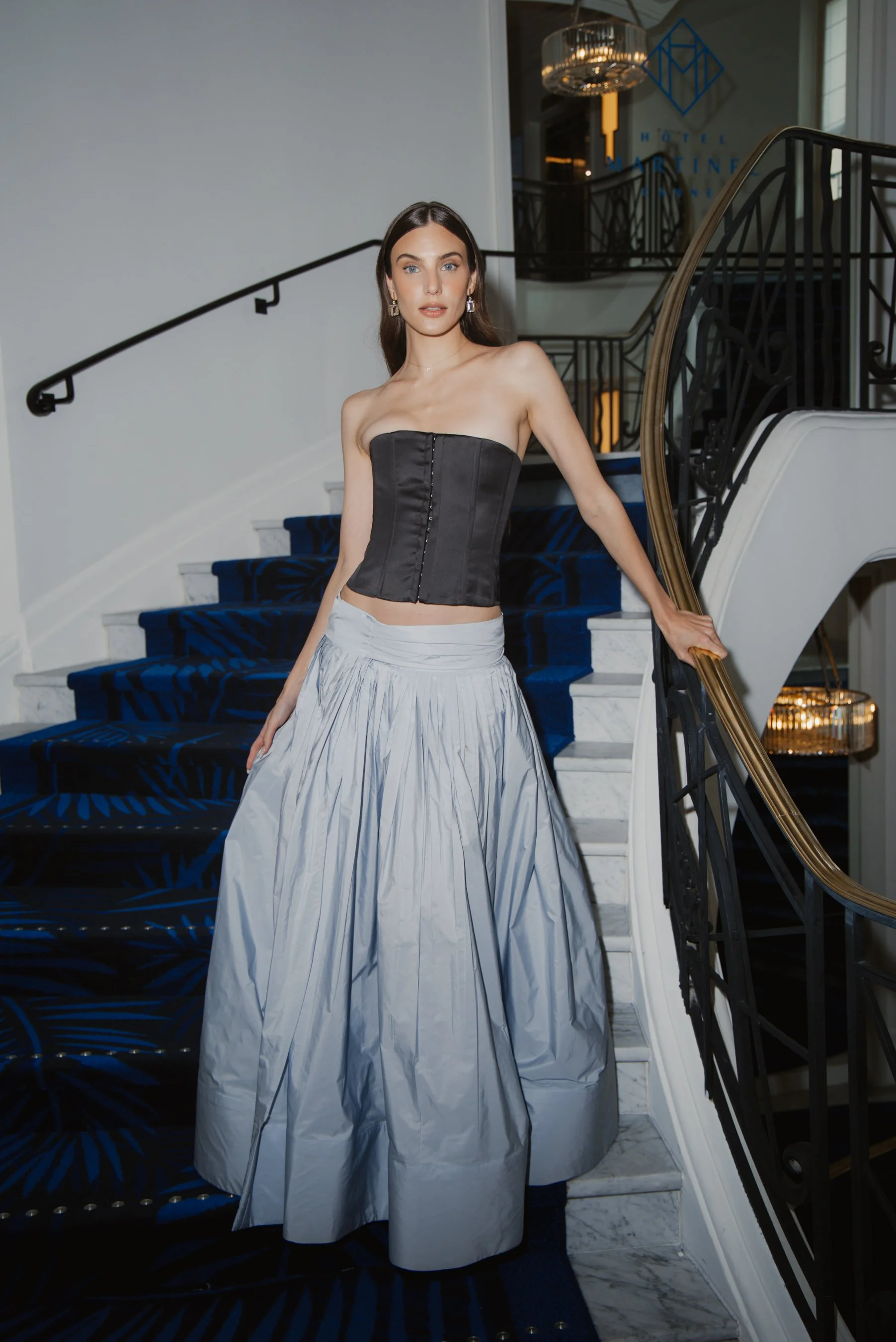 A woman in a strapless black bustier top and a long silver metallic skirt standing on a staircase in a hotel lobby.
