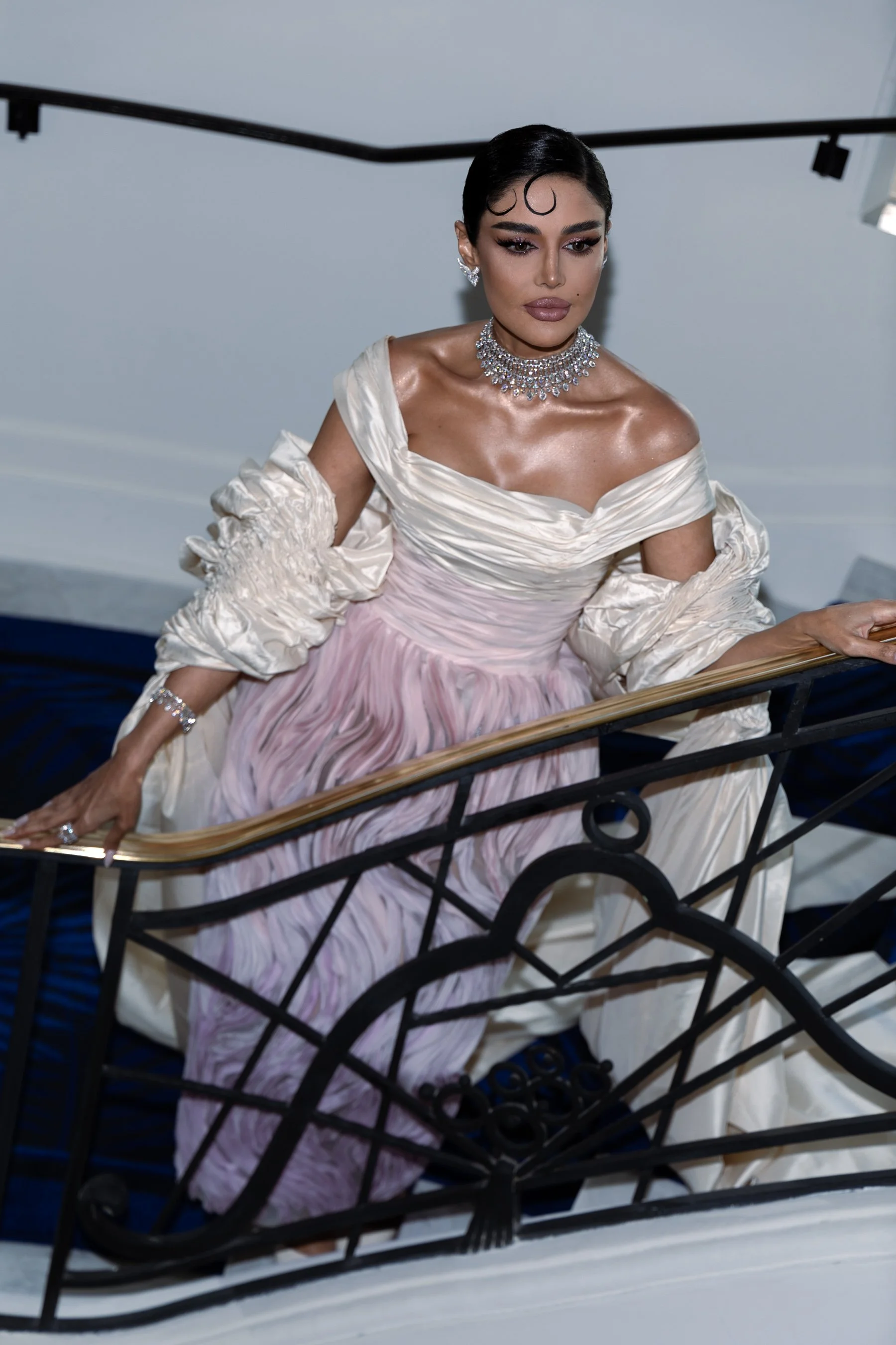 Woman in an elegant, off-the-shoulder gown with jewelry, standing on a staircase with a decorative black iron railing.