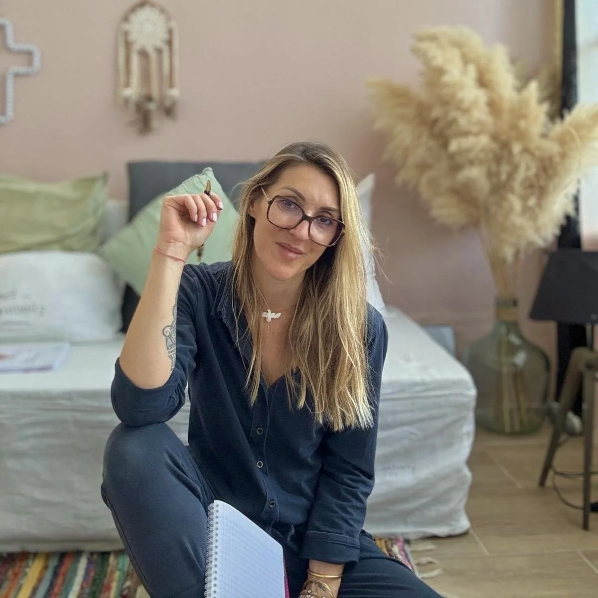 Une femme portant des lunettes assise au sol dans une chambre, tenant un stylo et un carnet, avec un lit en arrière-plan et un grand vase avec des plantes décoratives.