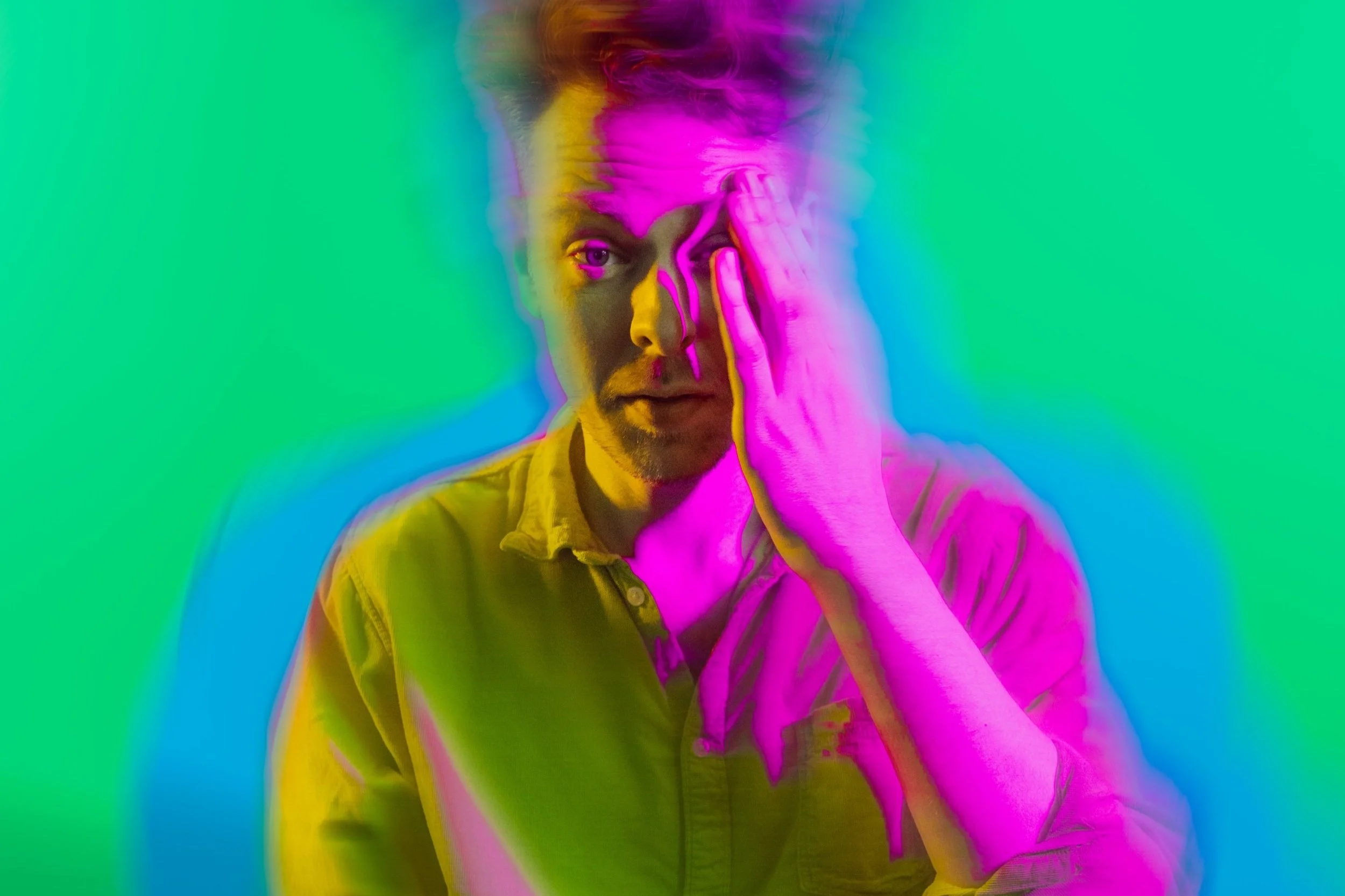 A person with one hand covering their face, surrounded by colorful neon effects, wearing a shirt, against a vibrant green and blue background.