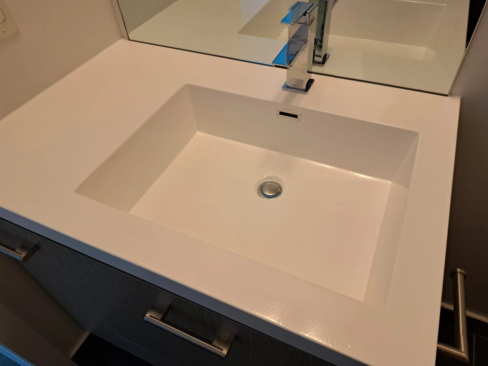 A modern white bathroom sink with a rectangular shape, a metallic faucet, a mirror above, and a small storage compartment beneath.
