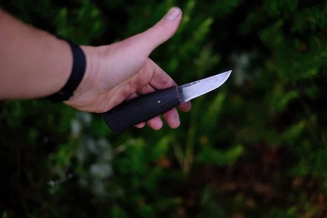Person holding a small knife outdoors against a background of green leaves.