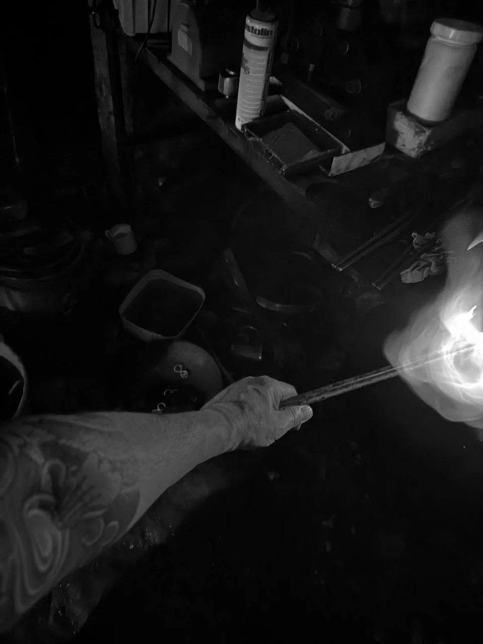 A person with a tattooed arm holding a lit torch or flame in a dark garage or workshop filled with tools, containers, and equipment.