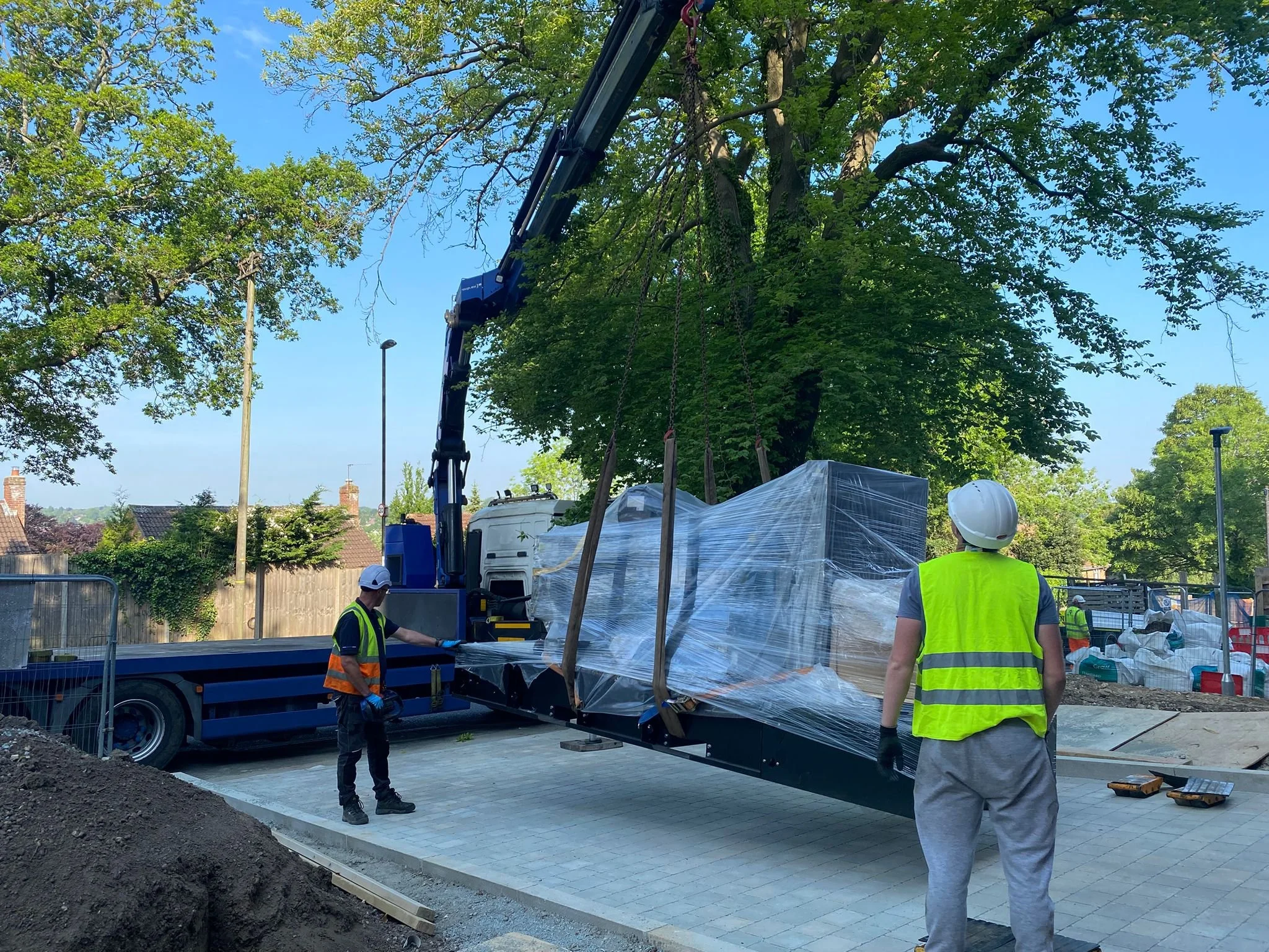 Construction workers in safety vests and helmets using a crane to lift a wrapped large object at a construction site with trees and a blue sky in the background. Brand New Diesel Generator