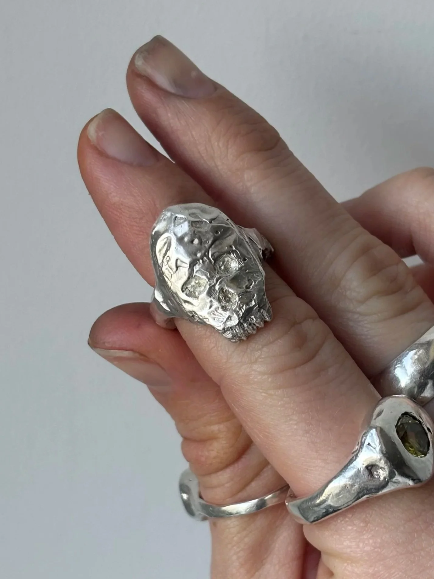 Close-up of a person's hand wearing silver jewelry, including a skull-shaped ring and a chunky bracelet, with a plain light background.