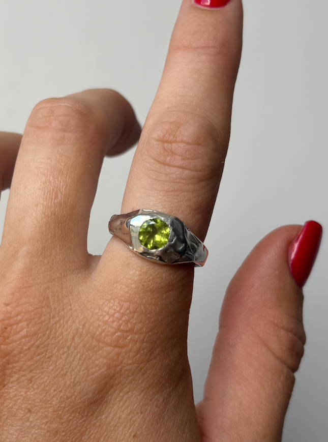 A hand wearing a silver ring with a round green gemstone on the middle finger. The hand has red nail polish and is set against a plain gray background.
