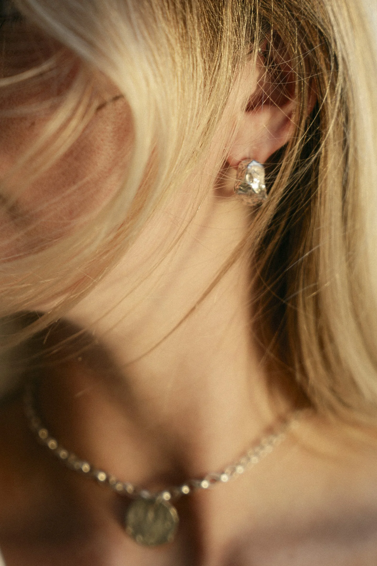 Close-up of a woman's ear with a sparkly earring, a pearl necklace, and blond hair.