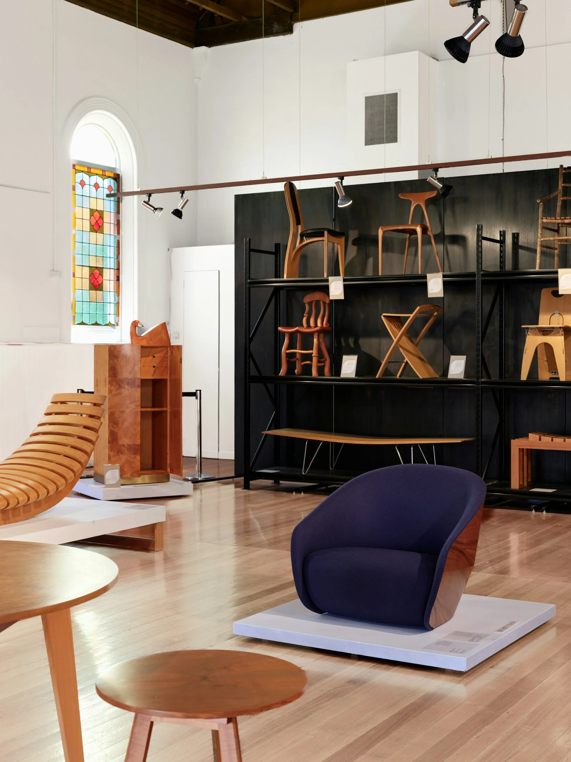 Binnenzicht van een museumruimte met houten en gestoffeerde stoelen en meubels, een gekleurd glas-in-lood raam, houten vloer en witte muren.