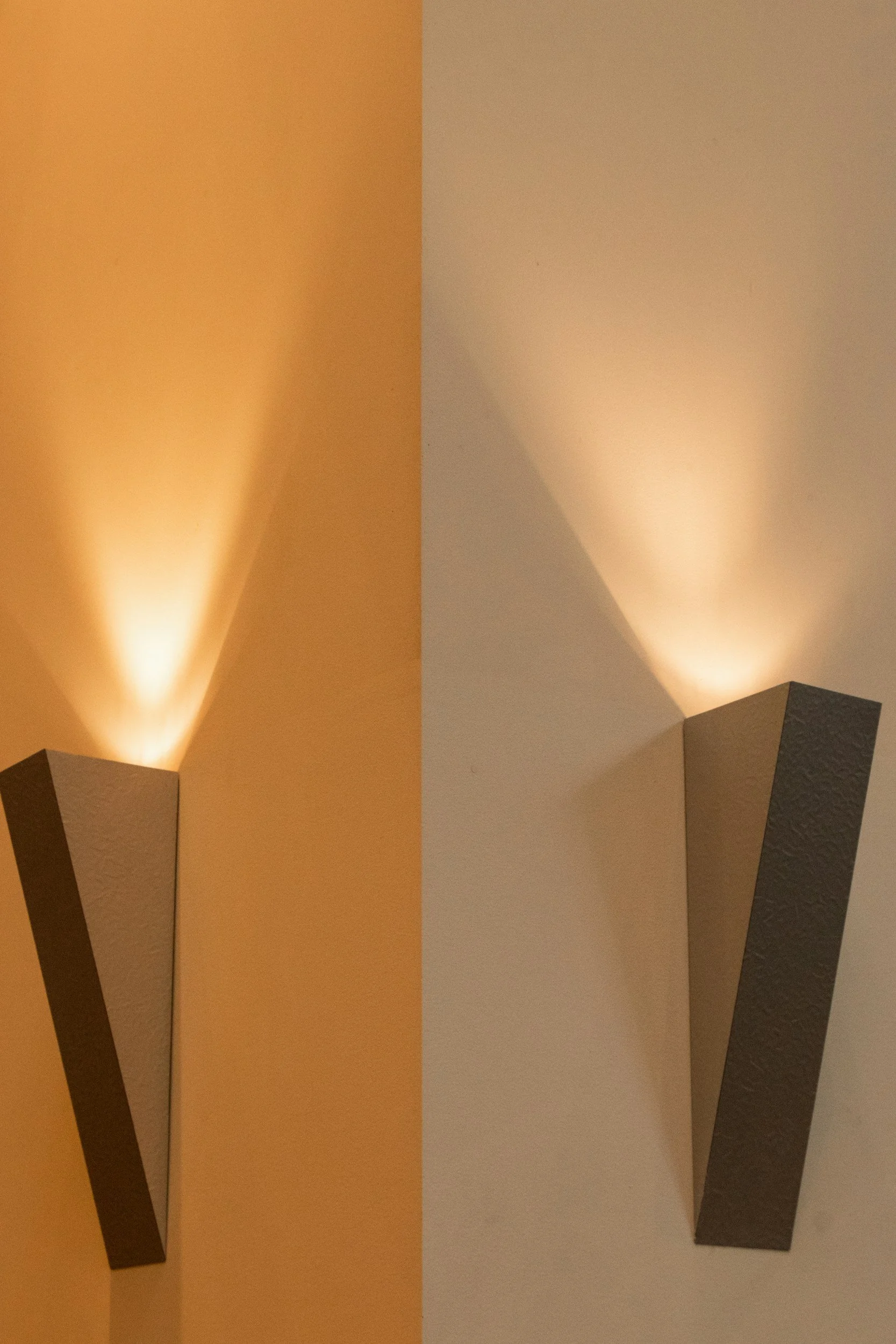 Twee muurlichten die licht naar boven uitstralen, één op een oranje muur en één op een witte muur.