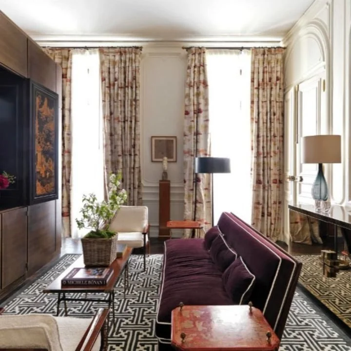 Een woonkamer met grote ramen met bloemige gordijnen, een paar zitbanken, een lage tafel met een plant, en een zijtafel met een lamp, ingericht in een elegant en stijlvol interieur.