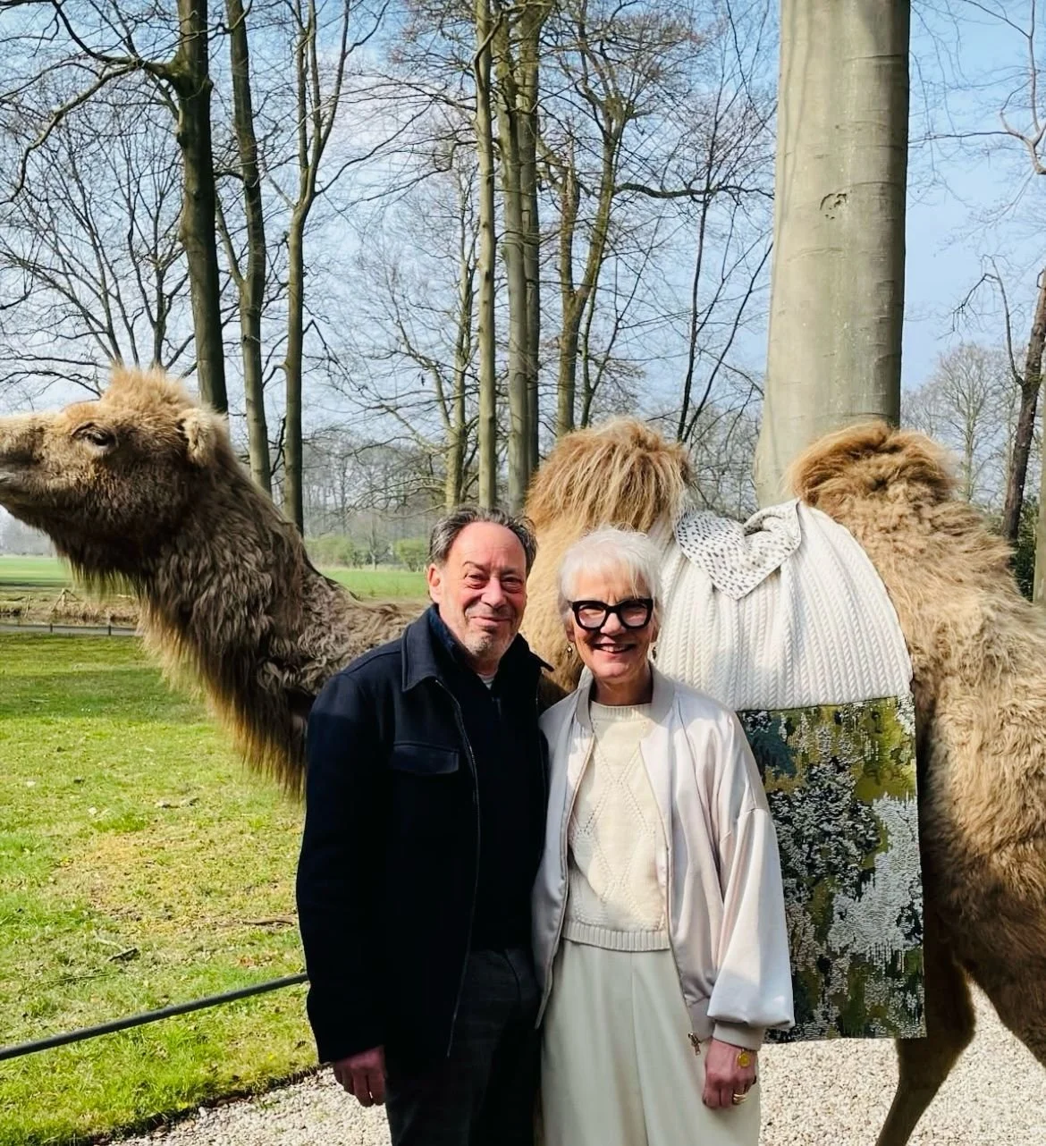 Twee mensen staan voor een kamelen in een park met bladaarbe bomen, de man en vrouw lachen en poseren voor de camera.