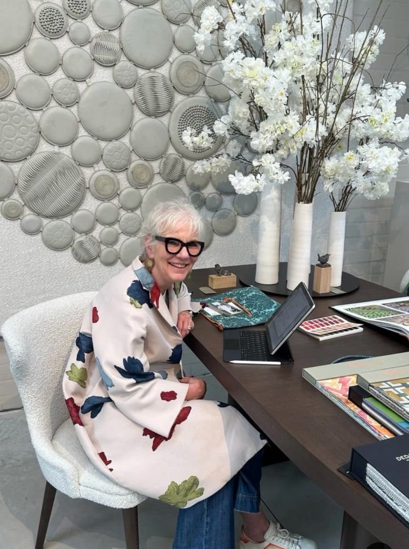 Een oudere vrouw met wit haar en grote zwarte bril zit aan een houten tafel, ze glimlacht. Ze draagt een witte jas met een bloemenpatroon. Op de tafel liggen een laptop, boeken, en ontwerpmappen. Achter haar hangen grote witte vazen met witte bloemen en takken, en op de muur hangt een kunstwerk van diverse ronde, textuurrijke objecten.